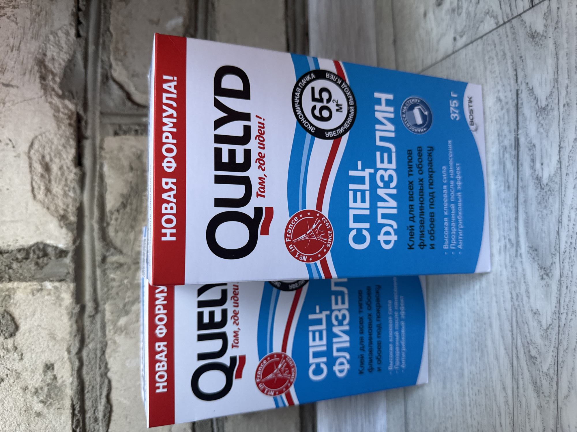 This is a close-up, overhead shot of two boxes of "QUELYD" brand wallpaper paste, specifically a "SPECIAL FLEESLIN" (СПЕЦ-ФЛИЗЕЛИН) product. The boxes are stacked vertically, with the top one partially obscuring the bottom one. The packaging is predominantly white and blue with red accents.
Prominently displayed on the front of each box is the brand name "QUELYD" in large, bold, black lettering. The top box features the text "НОВАЯ ФОРМУЛА!" (NEW FORMULA!) in red text. Both boxes highlight key features such as "65 м²" (65 m²) indicating coverage, and mention "Клей для всех типов флизелиновых обоев и обоев под покраску" (Glue for all types of non-woven wallpaper and wallpaper for painting). Additional benefits listed include "Высокая клеевая сила" (High adhesive strength), "Прозрачный после нанесения" (Transparent after application), and "Антигрибковый эффект" (Anti-fungal effect). A circular emblem with "France SINCE 1922" is also visible. The weight of the product, "375 г", and the brand "BOSTIK" are also present on the packaging.
The boxes are placed on a surface that appears to be a textured, grey brick wall or similar material, providing a rustic backdrop. The lighting suggests it might be indoors or under diffused daylight, with no strong shadows indicating a specific time of day. The scene is static, focusing solely on the product packaging. The location context provided is "Яромінскі сельскі Савет, Belarus".
