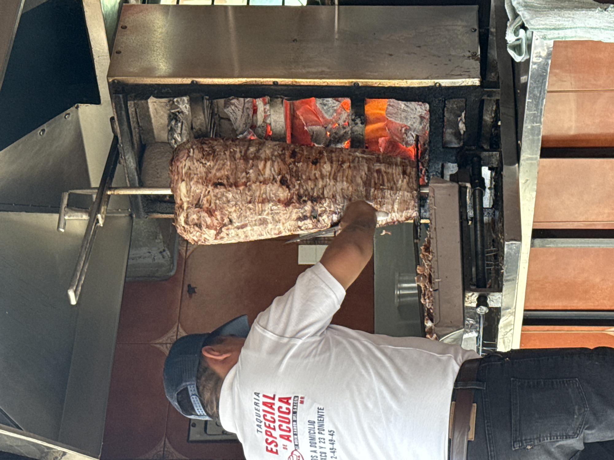 An adult male, viewed from behind, is actively slicing al pastor meat from a large, vertical trompo rotating over glowing red charcoal briquettes. The individual wears a dark blue baseball cap, a white short-sleeved polo shirt, and dark jeans with a brown belt. The polo shirt features red and black text, identifying the establishment as "TAQUERIA ESPECIAL DE ACUCA" and providing the address "FRANCISCO Y 23 PONIENTE" in Puebla, Mexico, along with a phone number. The sliced meat collects in a metal tray below the trompo. The scene is set within a commercial food preparation area, characterized by reddish-brown tiled walls with a faint decorative pattern, a stainless steel counter, and a metal exhaust hood above. A striped green and white towel rests on the counter to the right.