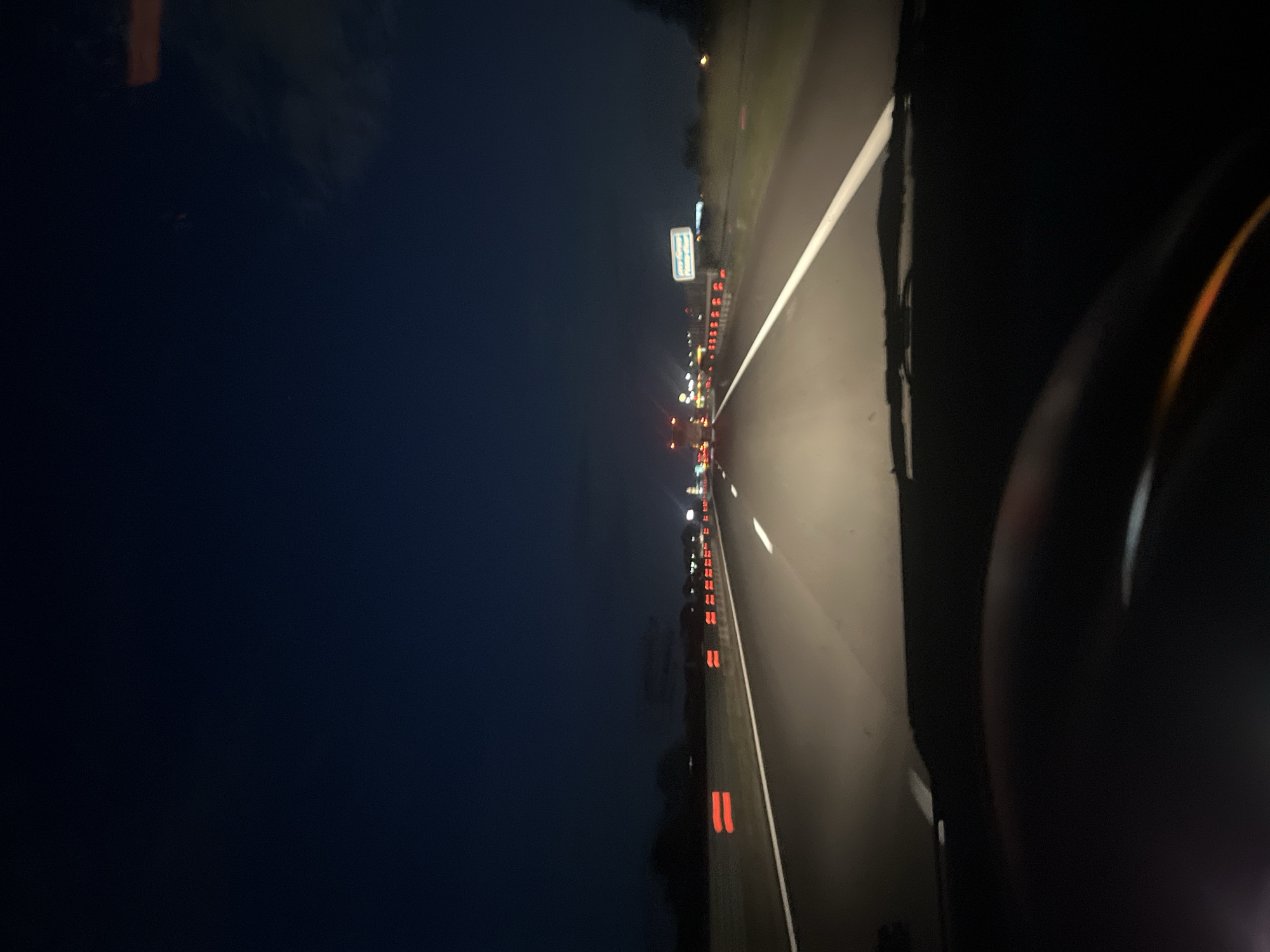 The image shows a view from inside a car driving on a highway at night. The highway has multiple lanes and is illuminated by streetlights and car headlights. There is traffic in both directions. The environment is dark and the sky is clear. The location is Unknown, Unknown.