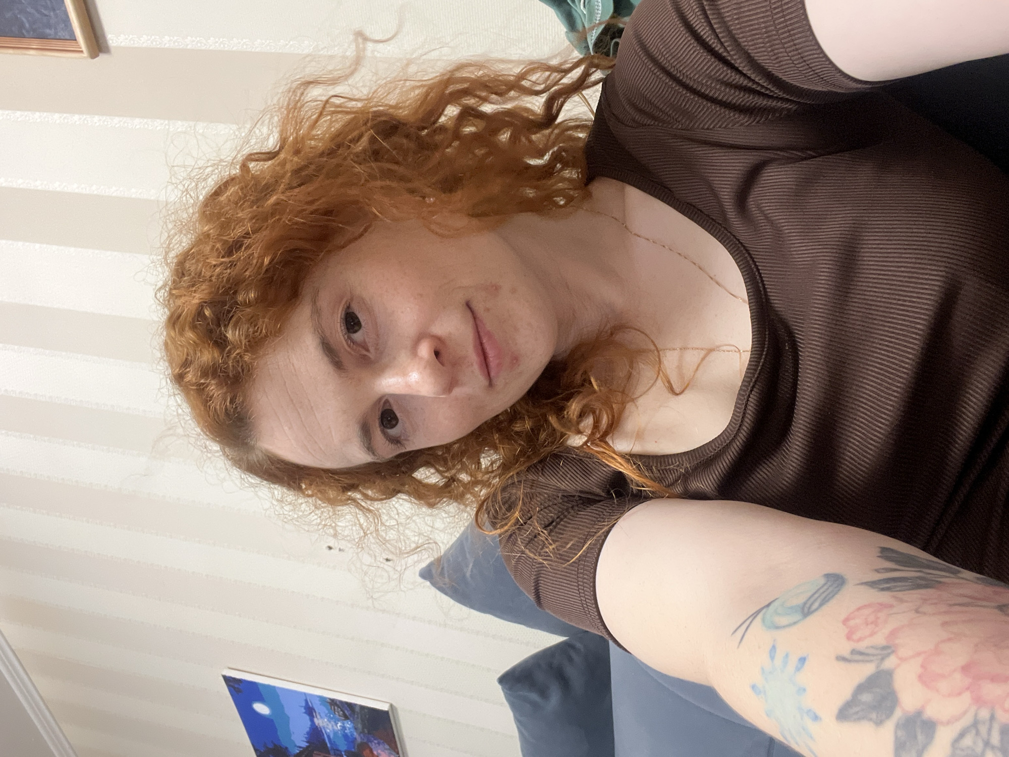 A person with red curly hair, light skin, and a brown top is pictured in a selfie. The person has a tattoo on their arm. Behind the person is a wall with vertical stripes and a picture frame hanging low on the wall. There is also a blue pillow visible. The location is Chaikovsky, Russia.
