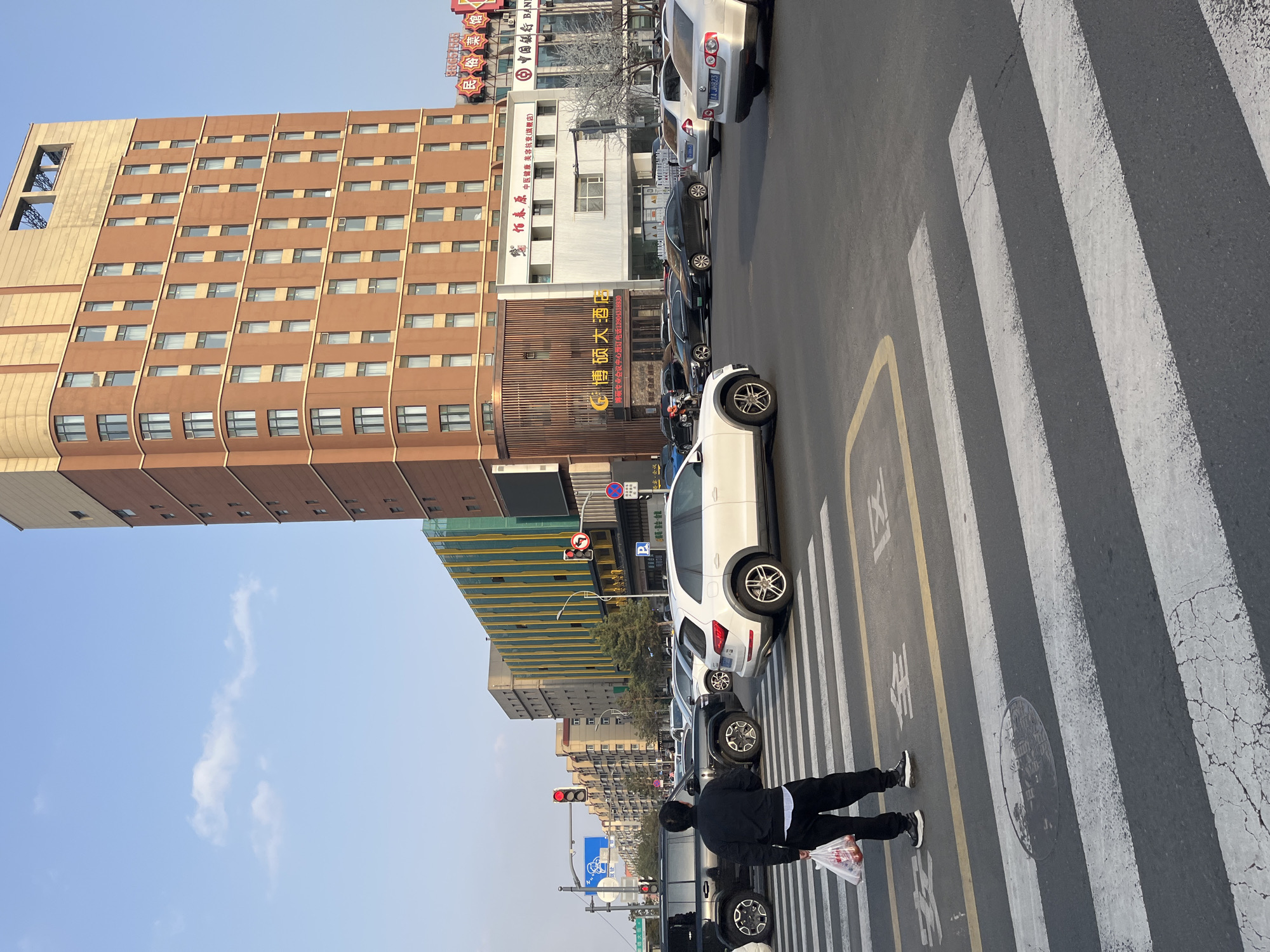 The image captures a vibrant urban street scene in Changchun City, China, on a clear, sunny day. The perspective is looking across a street with a pedestrian crosswalk in the foreground.
In the immediate foreground, a person is seen from behind, walking across the zebra crossing, holding a plastic bag containing items. They are dressed in a dark jacket, white top, and dark pants with white sneakers. The crosswalk markings include both white stripes and yellow lines, with Chinese characters for "安全通行" (Safe Passage) painted on the road.
The street is lined with parked cars, including several white and dark-colored vehicles. A white SUV with a license plate "BA JH823" is prominent on the right side of the crosswalk. In the background, a row of multi-story buildings dominates the skyline.
Notable buildings include a large, brown building with numerous windows and a distinctive curved base on the left, possibly a hotel. Above it, a red sign reads "博硕大酒店" (Boshuo Hotel) and below it, "博硕专业会议中心预订电话13904318930" (Boshuo Professional Conference Center Reservation Phone 13904318930). Further down the street, another building displays signs for "中国银行 BANK" (Bank of China) and "柏春园中医健康美容抗衰(旗舰店)" (Baichunyuan Traditional Chinese Medicine Health Beauty Anti-aging (Flagship Store)). Another building with a golden-toned glass facade is visible on the right.
Traffic lights and street signs are also visible, contributing to the urban setting. The sky is bright blue with a few wispy clouds.