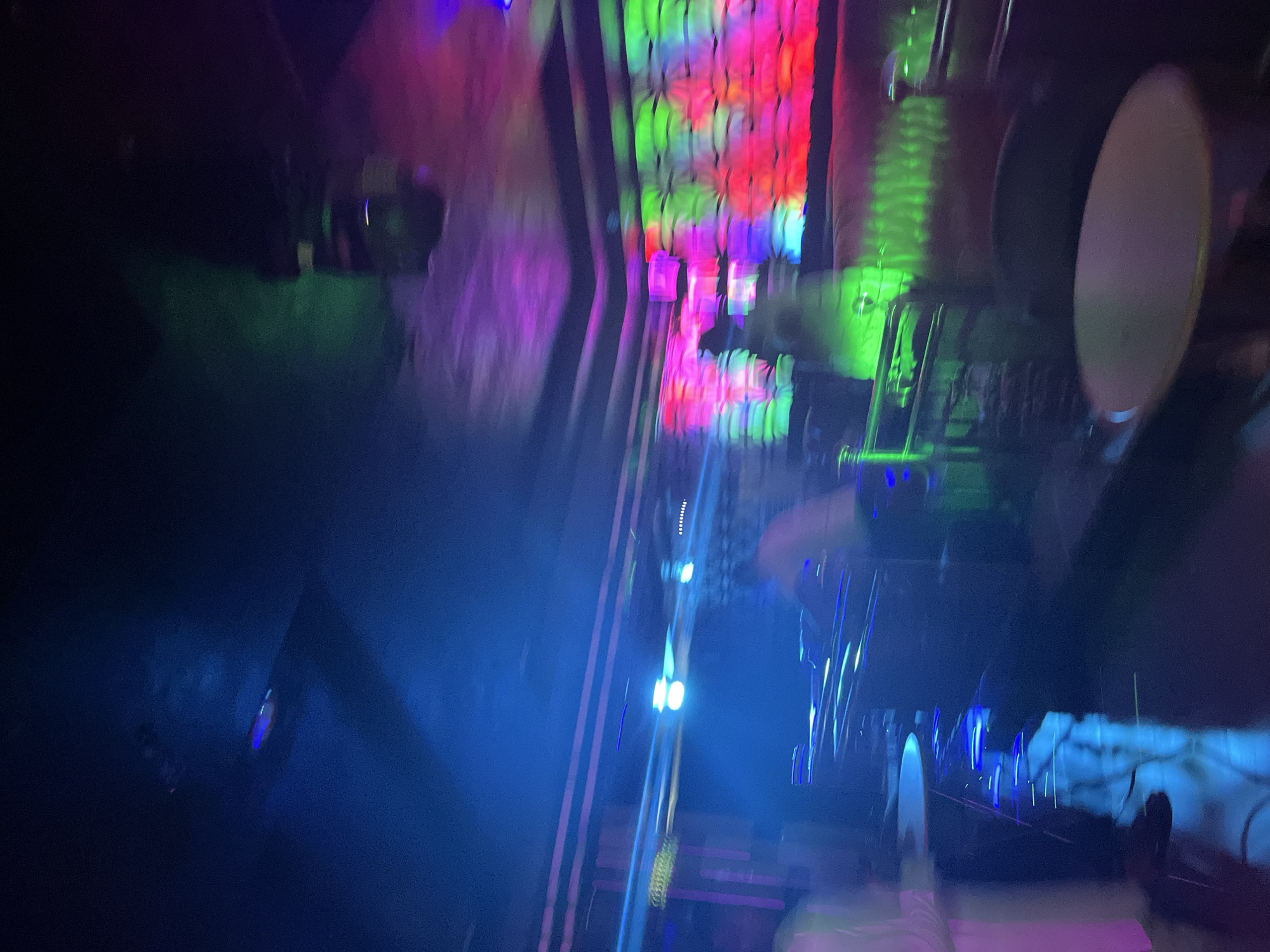 The image depicts the interior of a dimly lit nightclub with vibrant, colorful lighting. A DJ booth with illuminated equipment is visible, and several blurred figures are present, suggesting a lively atmosphere. The walls and ceiling are adorned with multi-colored lights that cast a dynamic glow throughout the space. Tables and seating are sparsely visible in the foreground and background. The overall impression is that of a bustling nightlife venue.