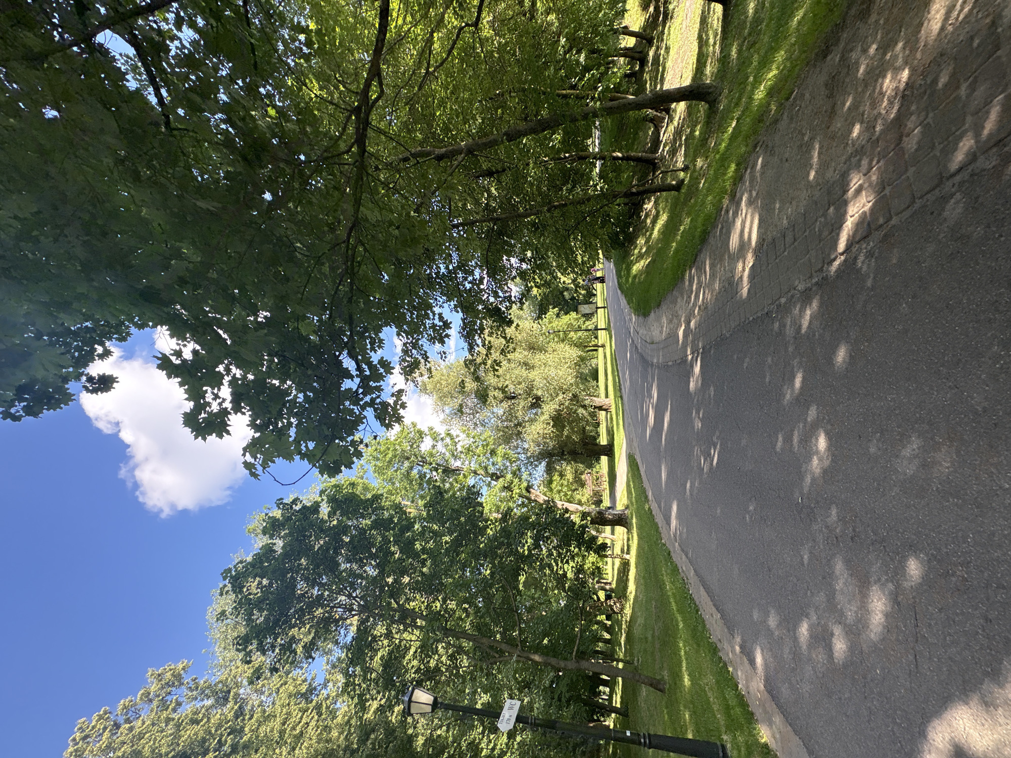 The image depicts a tree-lined pathway in a park setting. The pathway appears to be paved with asphalt and bordered by a brick edge on one side. Lush green grass and mature trees with dense foliage flank the path. Sunlight filters through the leaves, casting dappled shadows on the ground. The sky is blue with a few white clouds. A lamppost with a sign is visible on the left side of the path. In the distance, a few people can be seen walking along the path, and a wooden bench is nestled among the trees. The scene suggests a pleasant day for outdoor activity, likely in the late morning or early afternoon given the quality of light and shadows. The overall mood is serene and peaceful. The sign on the lamppost has some text, but it is illegible. The location is identified as Minsk, Belarus.