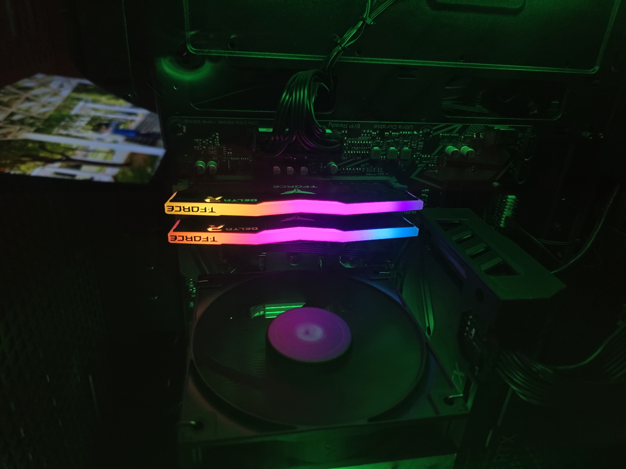 The image shows the interior of a computer case, focusing on the components related to cooling and memory. Two sticks of T-FORCE Delta RAM are prominently displayed, illuminated with RGB lighting showing a rainbow spectrum. Below the RAM is a CPU cooler with a large circular fan. The fan blades are dark, with a central area emitting pink and green light. The motherboard is visible, with various electronic components. Black cables are also visible and situated around the motherboard. The image is dimly lit, with a green cast dominating the scene. No specific location in Burgas, Bulgaria can be derived from the image.