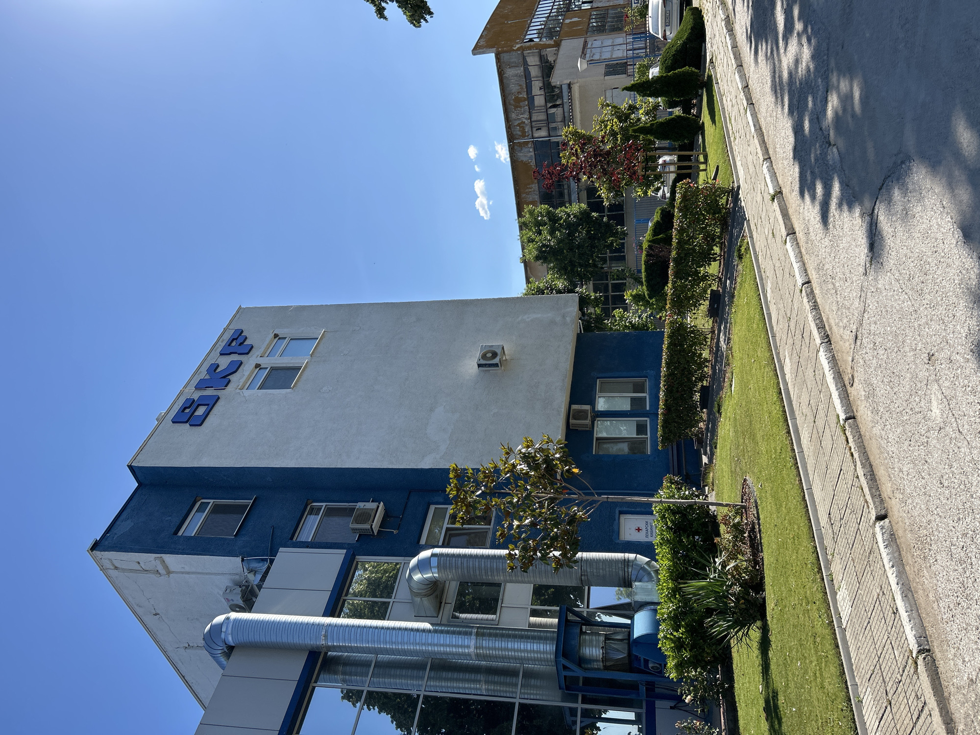 The image depicts the exterior of the SKF factory in Sopot, Bulgaria, on a clear, sunny day. The main building on the left is a two-story structure with a white upper section and a blue lower section. Large, reflective windows are visible on the lower part of the building, showing reflections of trees. Prominent blue "SKF" lettering is mounted on the upper white facade. A large, silver ventilation pipe runs vertically along the side of the building, attached to external ductwork. Several air conditioning units are mounted on the exterior walls. Green grass and manicured hedges form the landscaping in front of the building. A small tree stands in the middle of the lawn. A white door with a red cross symbol and text in Cyrillic script is also visible near the base of the blue section.
In the background, another industrial building with a weathered facade and large windows is visible. Trees of varying sizes and foliage are scattered throughout the scene. A sidewalk paved with square concrete tiles runs along the front of the buildings, adjacent to a grey asphalt road. Shadows of trees are cast across the road and sidewalk, indicating a bright sun. Two white cars are parked along the side of the road. The sky is a bright, clear blue with a few wispy clouds.