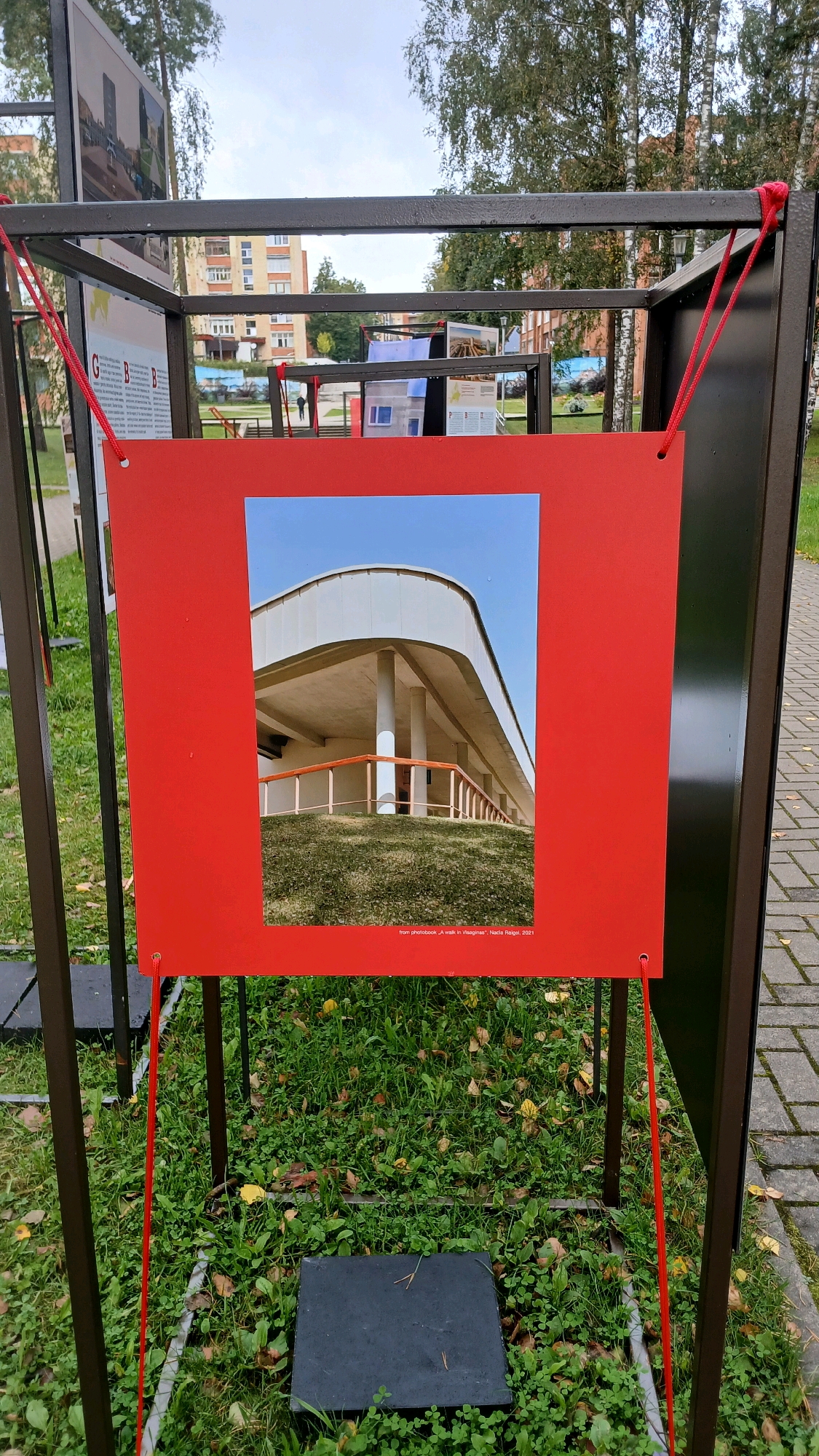 An outdoor art exhibition features photographs displayed on metal frames set on a grassy area next to a paved walkway. The central focus is a large red mat with a square opening, revealing a photograph of a modern building with a curved facade, supported by pillars, and a green, grassy embankment. The photograph's caption reads "from photobook „A walk in Visaginas“, Nadie Reigll, 2021." In the background, apartment buildings and trees are visible, suggesting an urban setting. Red ropes are used to hang some of the display elements. The weather appears overcast.