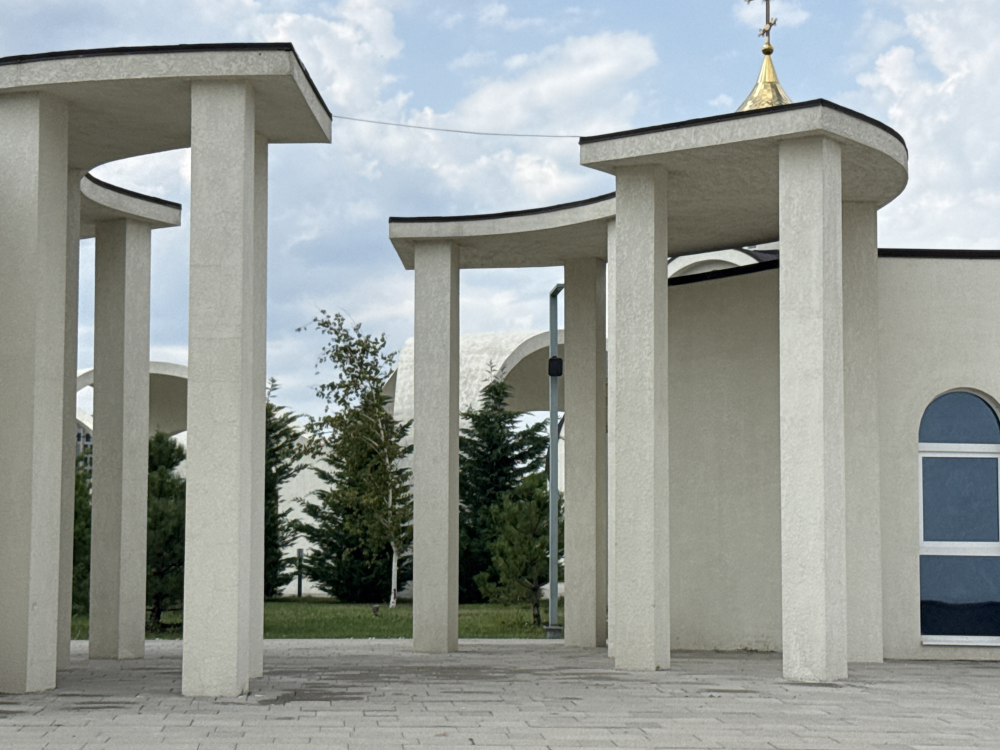 This is a medium shot, eye-level, outdoor photograph of a modern building with curved architectural elements and thick, light-colored pillars. The building appears to be a religious structure, possibly a church or chapel, indicated by the golden dome and cross visible on the roof. The building is off-white with a dark, flat roofline. To the right of the building, there is a tall, arched window with a dark blue pane and white trim. In the foreground, there is a paved walkway made of light gray rectangular stones. Behind the pillars and to the left, there are several evergreen trees and a deciduous tree with lighter bark, suggesting a well-maintained landscape. The sky is partly cloudy with a mix of white and gray clouds against a blue sky. The scene is static, with no people or animals visible. The lighting suggests it is daytime, possibly late afternoon or early morning due to the soft light. The architecture suggests a contemporary design.