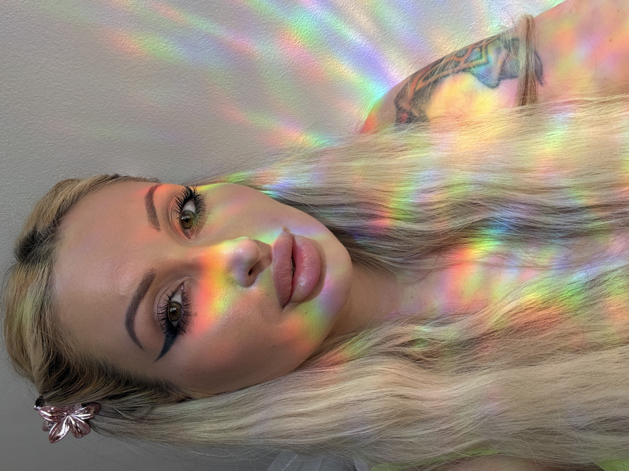A close-up, medium shot features a woman with blonde hair adorned with a pink flower hair clip. She has prominent makeup, including eyeliner and lipstick. Rainbow-colored light streaks across her face, neck, and shoulder, partially obscuring her skin and hair. A tattoo is visible on her upper arm. The background is a textured, off-white wall. The lighting creates a prism effect.
