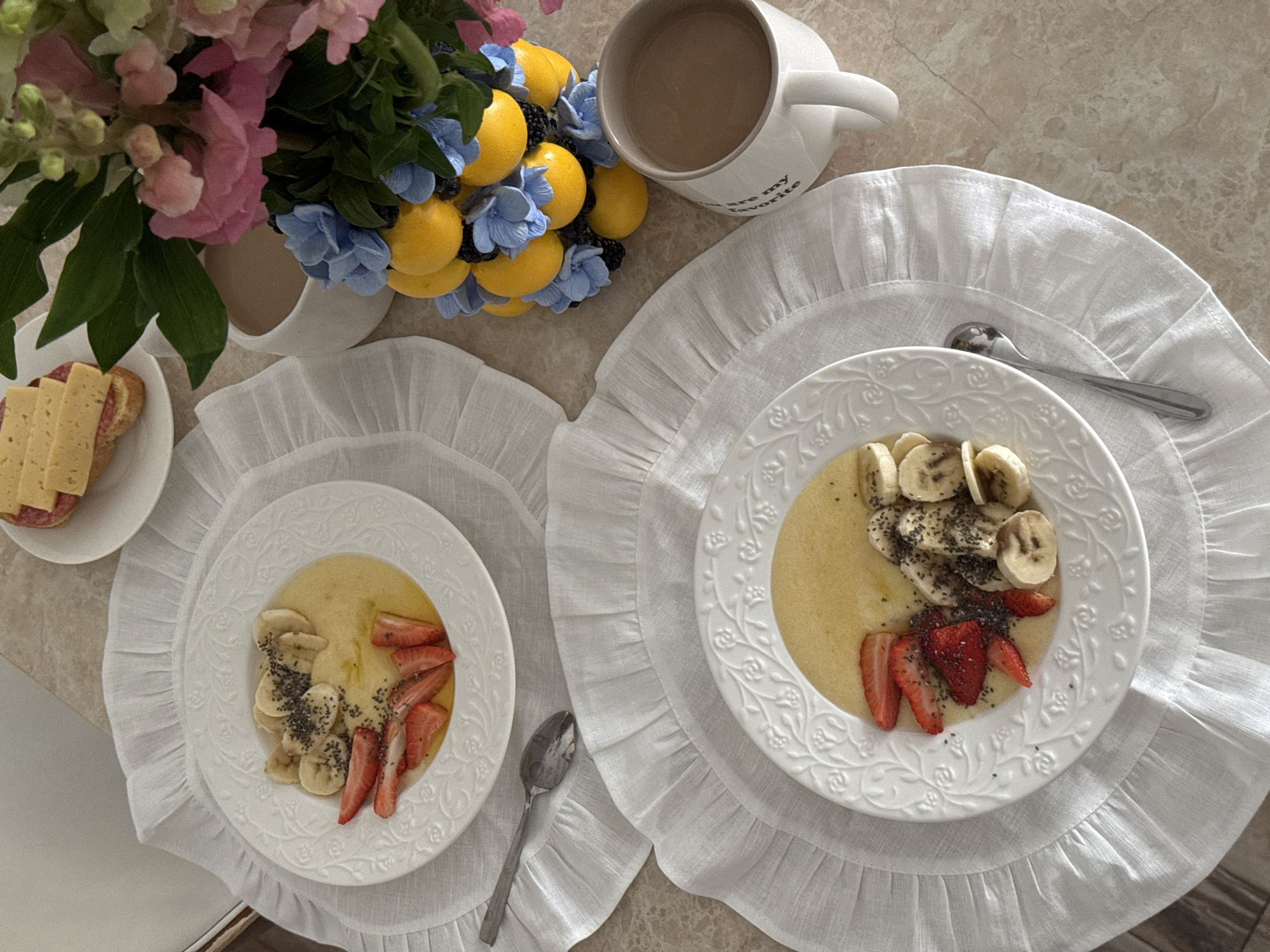 The scene depicts a table setting with two plates of breakfast cereal topped with sliced bananas and strawberries, sprinkled with chia seeds. Each plate rests on a white ruffled placemat. A spoon is placed beside each plate. In the background, there's a vase with flowers and a mug filled with liquid, possibly coffee or tea. A small plate with cheese and salami is also visible. The scene has a bright and airy feel, typical of a breakfast setting. The location is not possible to ascertain from the image.