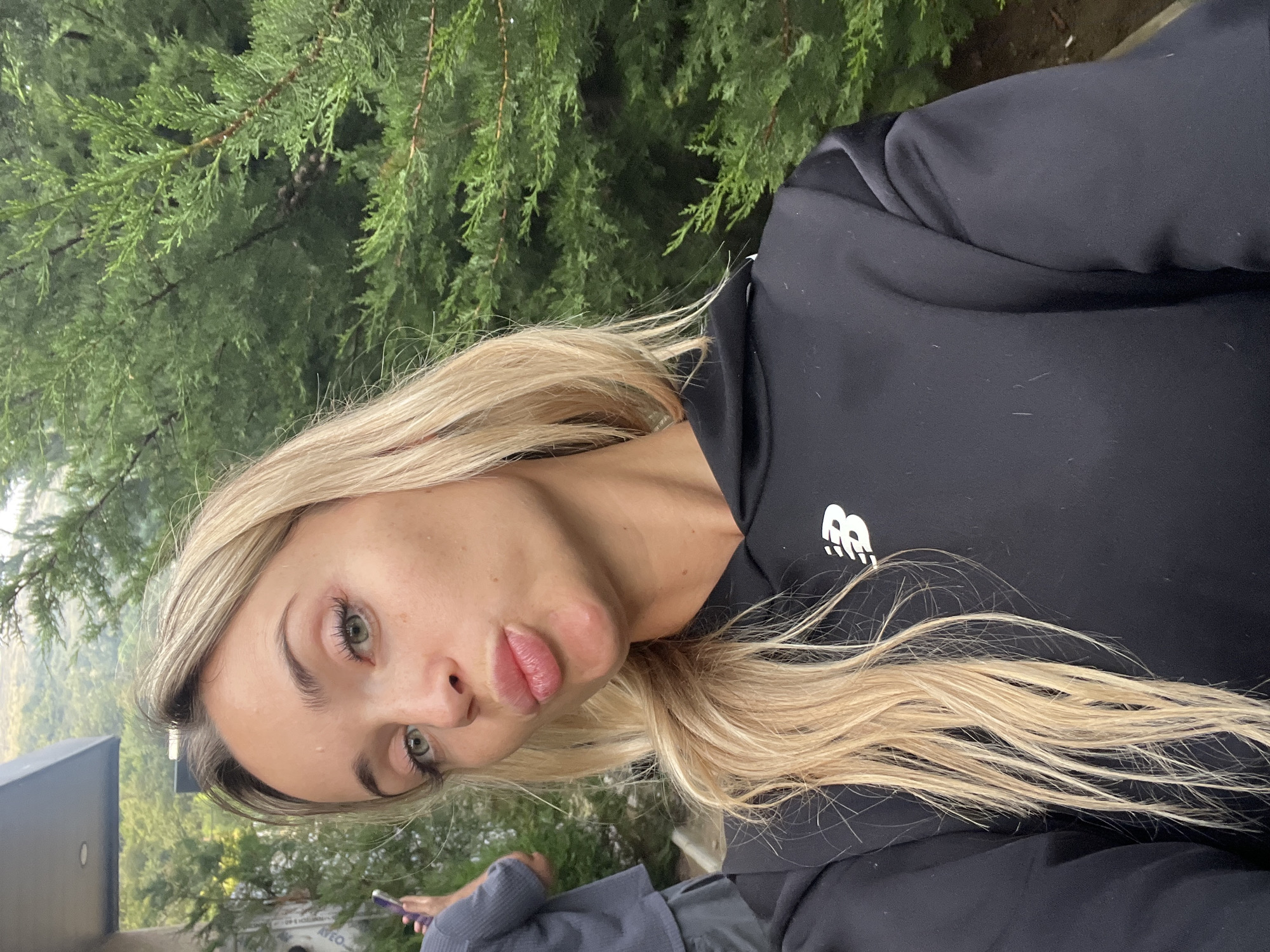A young blonde woman with green eyes and fair skin poses for a selfie outdoors, puckering her lips. She is wearing a black hooded sweatshirt with a white New Balance logo on the chest. Her blonde hair is long and falls over her shoulders. Behind her, a person in a gray shirt and dark shorts is partially visible, holding a mobile phone. The background features lush green trees and a hint of a mountainous landscape in the distance. The scene appears to be set in Tsodoreti, Georgia.
