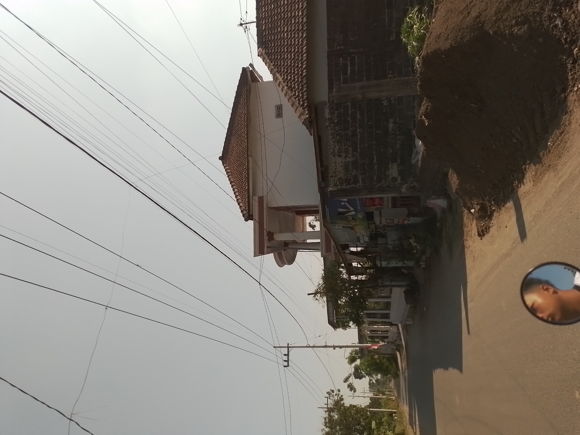 The image captures a street scene in Ngancar, Indonesia, likely during daytime due to the bright, clear sky. In the foreground, a section of an asphalt road is visible, with a shadow cast across it from an unseen object, possibly a vehicle. A motorcycle's side mirror is reflected in the lower center of the image, showing a partial view of a young man's face with short, dark hair.
The background features several houses. To the right, a white building with a terracotta tile roof stands prominently. Adjacent to it is another building with a similar tile roof and a dark brick wall. A large pile of dark brown soil is located in front of this building. Further down the road, on the left, a street with some trees and more houses can be seen. A utility pole with multiple wires stretches across the upper portion of the image, indicating the presence of electricity infrastructure. A small Indonesian flag is attached to a pole near the road on the left. The overall environment appears to be a residential or semi-rural area within the town of Ngancar.
