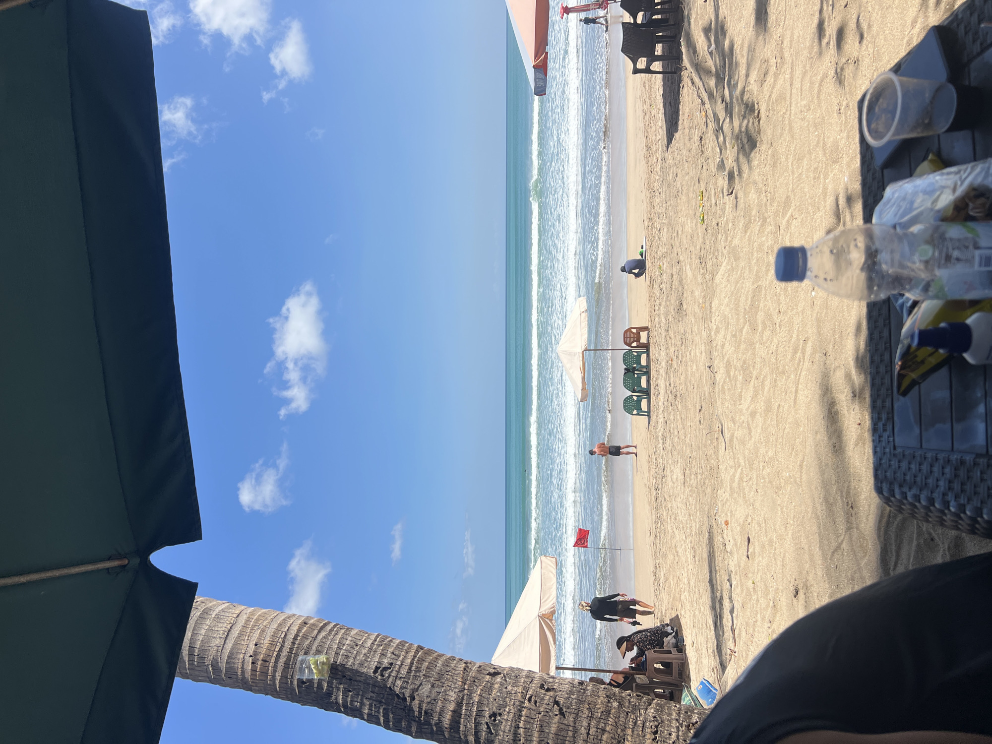 This is a photograph taken from a shaded seating area, looking out towards Kuta Beach, Indonesia. In the foreground, a black woven table holds a plastic bottle of water, a plastic cup with dark liquid, a phone, and a small blue bottle. The table is partially covered by shadows from a large dark green umbrella. To the left, a section of a person's dark clothing is visible.
The beach is composed of light-colored sand. Several beach umbrellas are set up, with two prominent beige umbrellas and one orange and white umbrella. A tall palm tree trunk is visible on the far left, with a clear plastic bag containing what appears to be green items attached to it.
On the sand, there are several people. Near the left beige umbrella, two individuals are seated on chairs. Further out on the sand, a man in a black long-sleeved shirt and shorts stands near a red flag planted in the sand. Another man in shorts is standing further out towards the water. In the middle ground, a person is sitting on the sand facing away from the camera. Several green and brown plastic chairs are arranged on the sand.
The ocean is a vibrant turquoise with white-capped waves rolling towards the shore. The sky is a clear bright blue with scattered white clouds. The overall impression is a sunny day at a popular beach in Kuta, Indonesia.