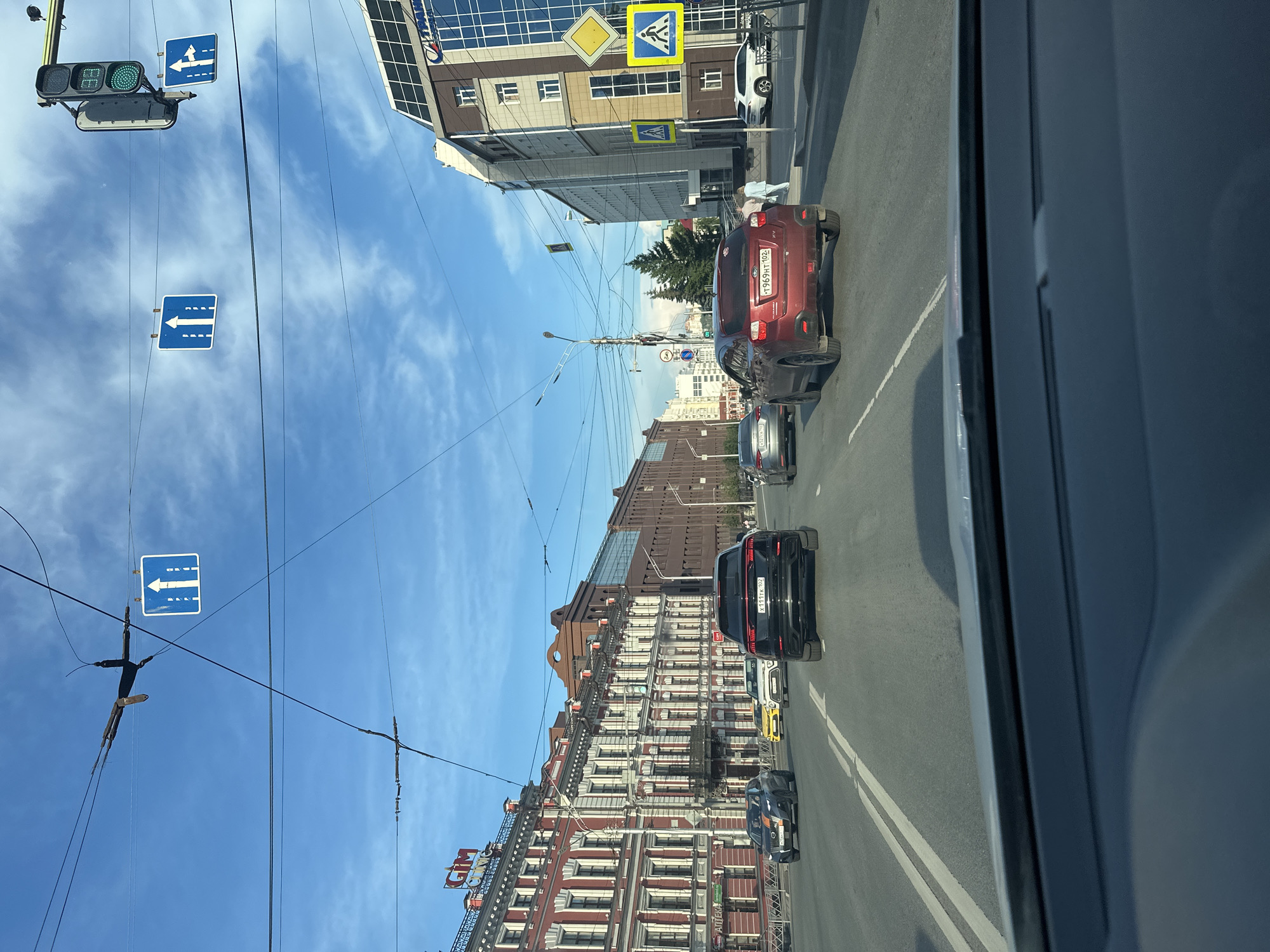 The image shows a street scene in Ufa, Russia, on a clear day with a blue sky and scattered clouds. A multi-lane road with white lane markings dominates the foreground and midground. Several cars are traveling on the road, including a black SUV in the immediate foreground, a red SUV to its right, and other vehicles further ahead. Pedestrian crossings with yellow and blue signage are visible on the right side of the road. Buildings line both sides of the street, with architectural styles varying from ornate, historic facades on the left to more modern glass and concrete structures on the right. Overhead, power lines and traffic lights are suspended across the road. A traffic light on the right shows a green signal. Road signs indicating lane directionality are also visible, suspended over the street. The scene depicts a typical urban traffic environment.