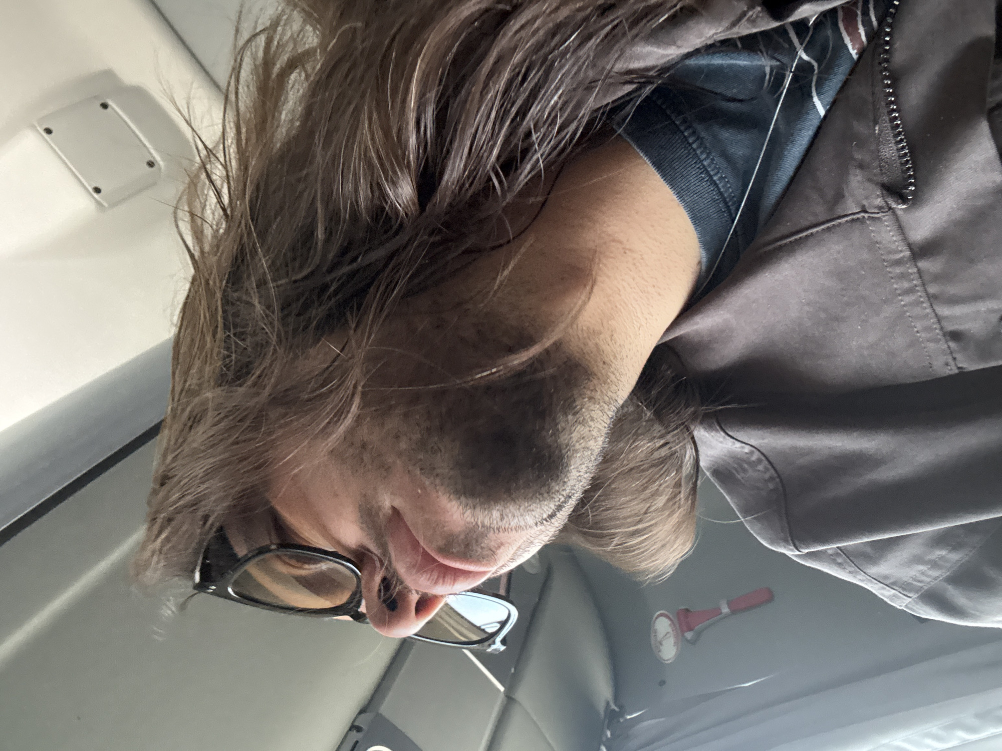 The image, which appears to be rotated 90 degrees clockwise from its original capture, depicts a close-up, low-angle selfie of a person, likely male, with long, wavy brown hair and stubble or a short beard. The individual is wearing dark-framed glasses, a dark t-shirt, and a dark grey or brown jacket with a prominent zipper. Their lips are slightly parted, and they appear to be looking downwards towards the camera with a neutral expression.
The setting is the interior of what seems to be a vehicle, possibly a car, truck, or bus, indicated by the light-colored ceiling and interior wall panels. In the upper left background, a white rectangular fixture with four dark screws is visible on the ceiling, potentially a light, sensor, or vent cover. Towards the lower right, mounted on a light wall, is a red object with a white circular base, resembling an emergency tool like a safety hammer or part of a fire extinguisher system. The white circular base has some indistinct red markings or text.
The perspective suggests the photo was taken from below the person, who is either leaning over or looking down at the camera. The lighting is diffused, consistent with an indoor setting, and does not indicate a specific time of day beyond being sufficiently lit. The location context for this media file is Valle de Chalco Solidaridad, Mexico. No clearly legible text is visible within the frame.