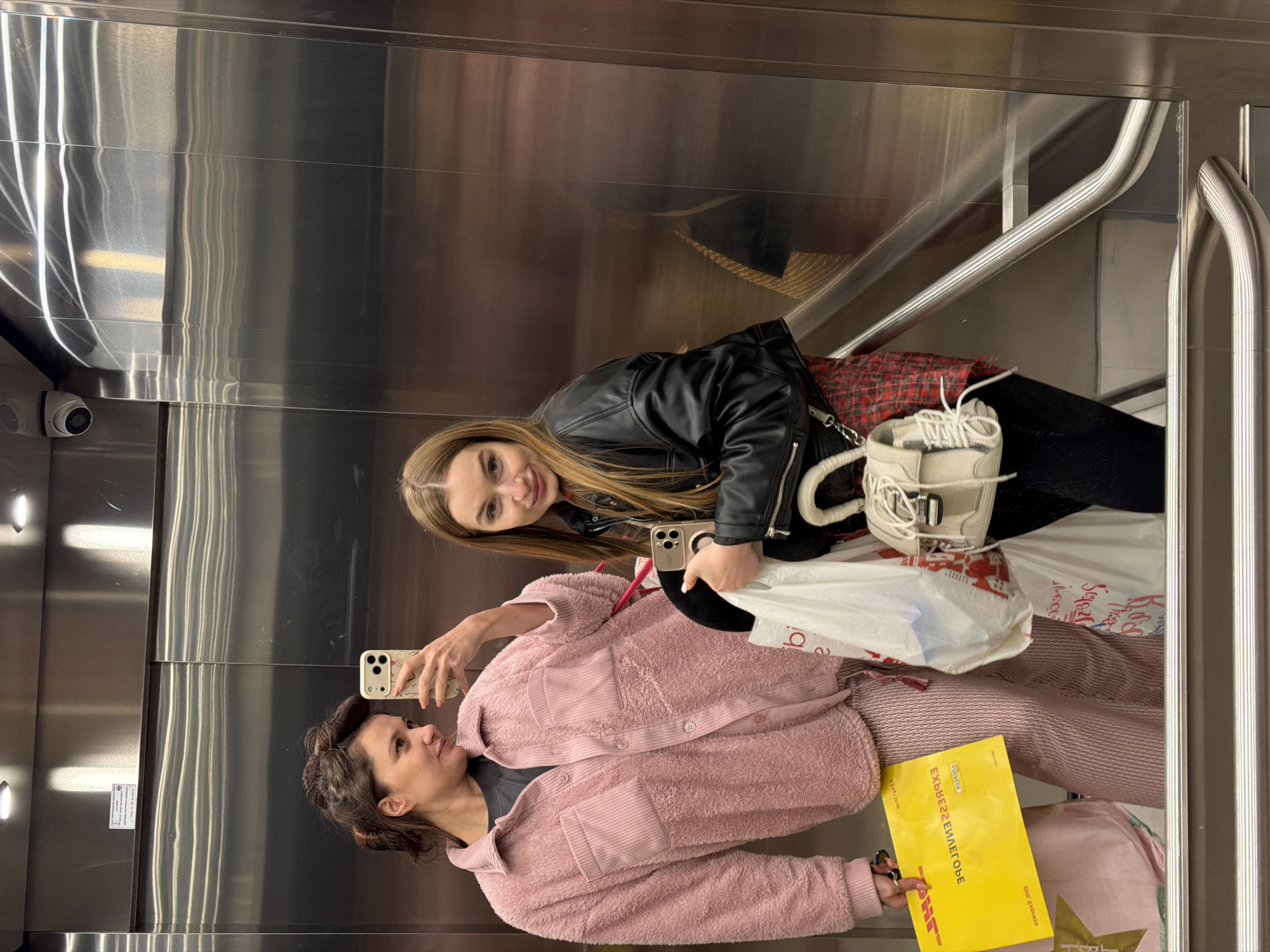 Two women take a mirror selfie inside an elevator. The taller woman, presumably younger, has long blonde hair and is wearing a black leather jacket. The shorter woman is wearing a pink jacket. She holds a phone in her hand and a yellow DHL package in the other. There are shopping bags in the elevator and what appears to be a handrail. A security camera is visible in the upper left corner. The location is Belgrade, Serbia.