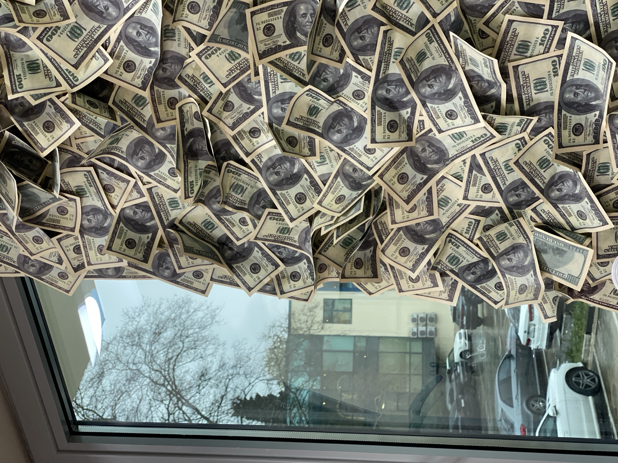 A window view in what appears to be Sochi, Russia, is partially obscured by a collection of one hundred U.S. dollar bills. The bills are arranged in a dense pile above the window frame. The window offers a blurred view of bare trees, a multi-story building with air conditioning units, and several parked cars.