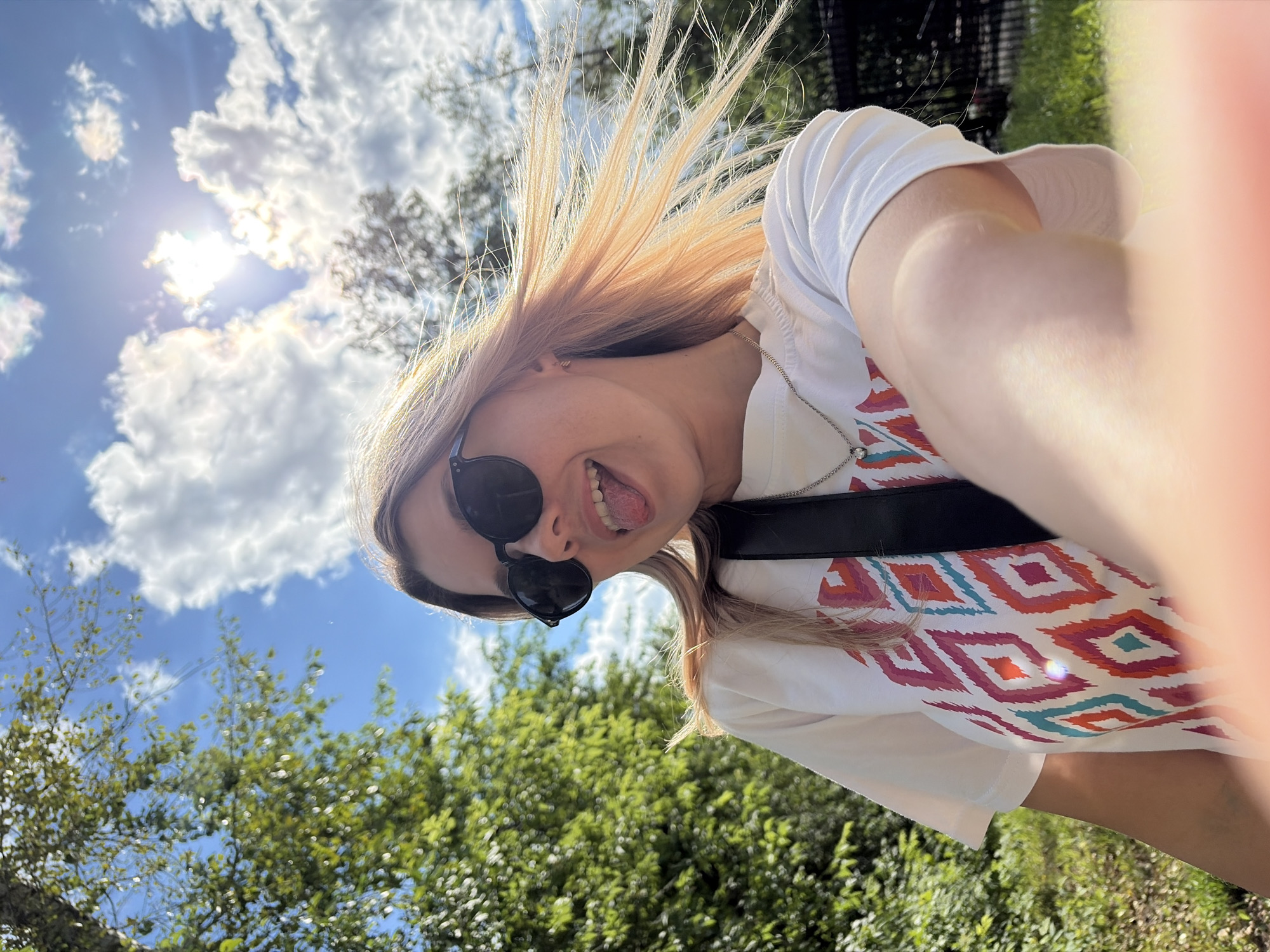 The image shows a young woman with blonde hair and sunglasses taking a selfie outdoors. She is wearing a white t-shirt with a colorful geometric pattern and a silver necklace. Her tongue is sticking out, and her hair is blowing in the wind. The background consists of green trees, a blue sky with white clouds, and sunlight filtering through. The scene appears to be in a park or a natural area. There are no explicit indicators of the specific location in Chaikovsky, Russia within the visual information provided.