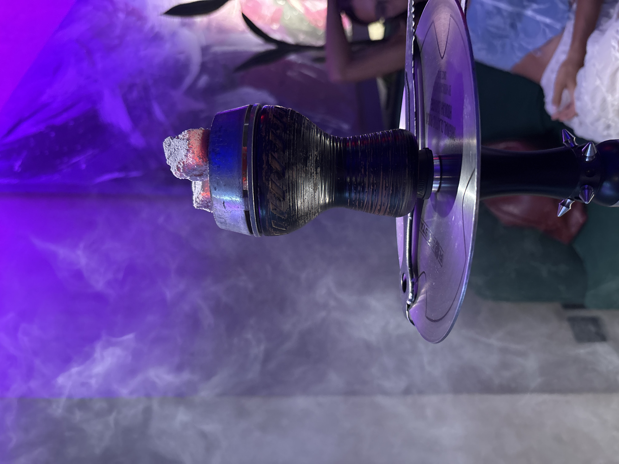 The image shows a close-up of a hookah. The bowl is loaded with burning coals. The hookah has a dark stem with a silver tray. Smoke fills the air. The location is Kuta Selatan, Indonesia. People are visible in the background.