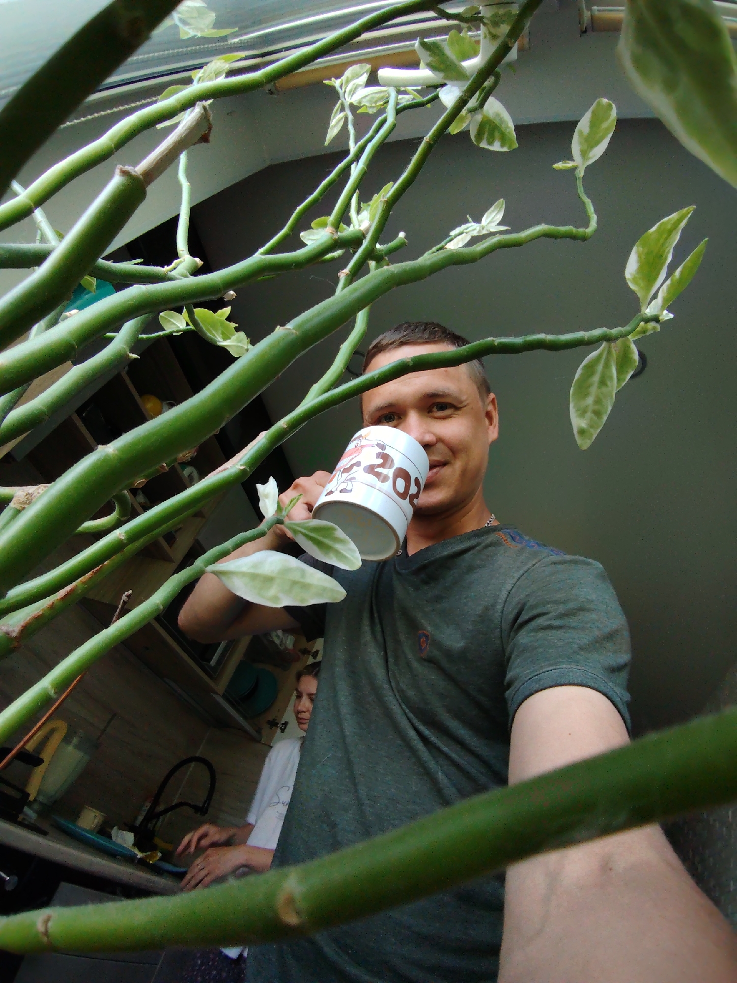 The image is taken indoors, likely in an apartment kitchen in Chaikovsky, Russia. A man is holding a mug with "202" visible on it, possibly drinking from it. He's wearing a dark green t-shirt. A woman is at the kitchen counter in the background, possibly doing dishes or food preparation. A kitchen sink, faucet, and some appliances are visible on the counter. The scene is partially obscured by a plant with green stems and variegated leaves. The lighting is a combination of natural and artificial.