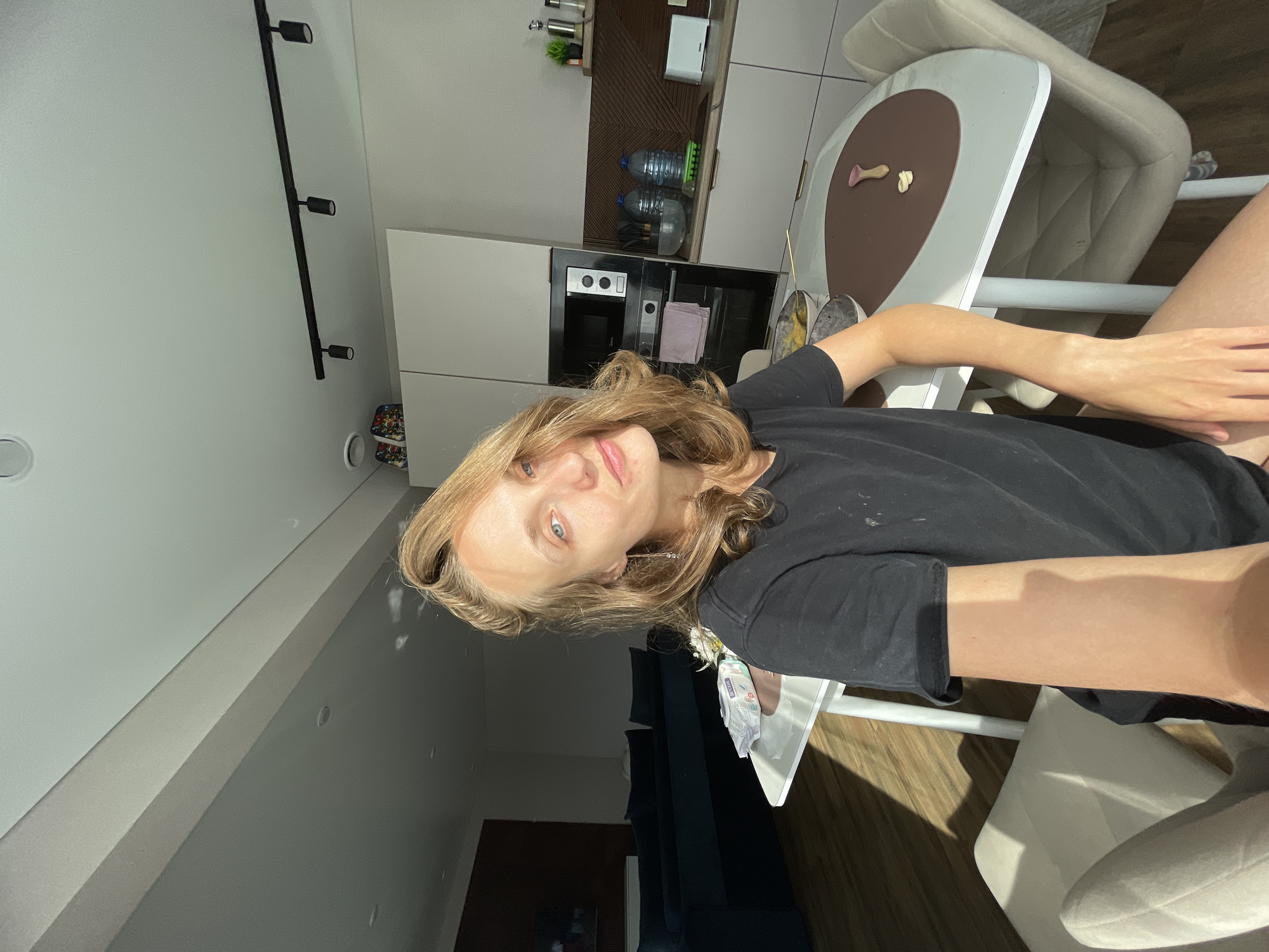 A young woman with blonde hair and blue eyes is sitting at a white oval table in a well-lit kitchen. She is wearing a black t-shirt. The kitchen has modern white cabinets, a built-in oven, and a countertop with bottles of water and a toaster. There is a window with sunlight streaming in, casting shadows on the floor and the woman's arm. In the background, a living room area with a dark blue sofa is visible. The overall impression is of a casual, everyday scene in a home. The location of Podshivalovo, Russia is not depicted in the image.