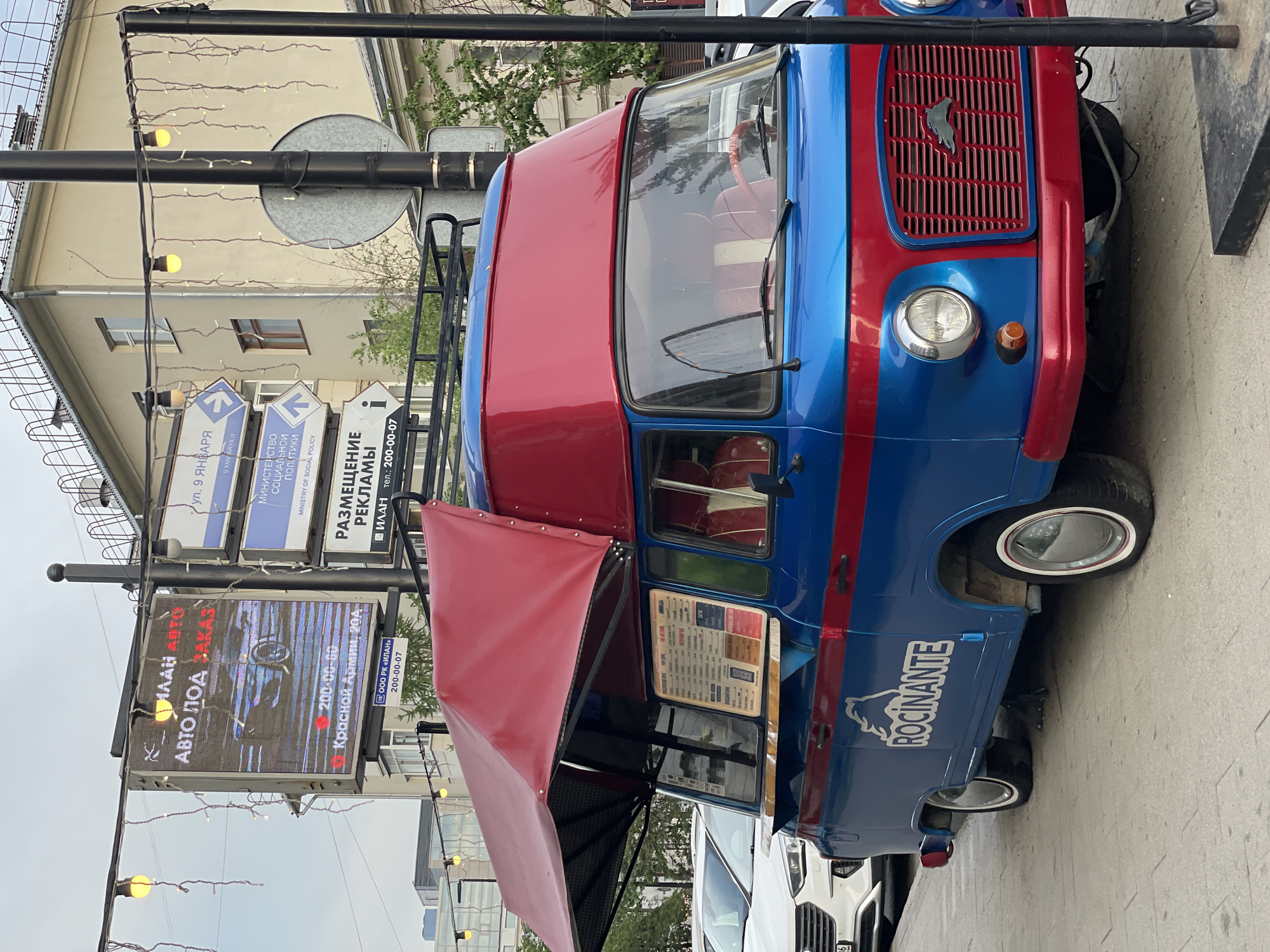 The image shows a bright blue and red food truck, identified by the name "ROCINANTE" on its side, parked on a sidewalk. The truck has a red awning extended over a service window displaying a menu. In the background, there are buildings, street signs, and other vehicles. One prominent street sign indicates "ул. 9 ЯНВАРЯ" (9 Yanvarya Street), suggesting the location is Krasnoyarsk, Russia. Another sign shows "Красной Армии, 20А" (Krasnoy Armii, 20A), which is a street in Krasnoyarsk. The overall scene depicts an urban street setting, likely during daytime with overcast skies.