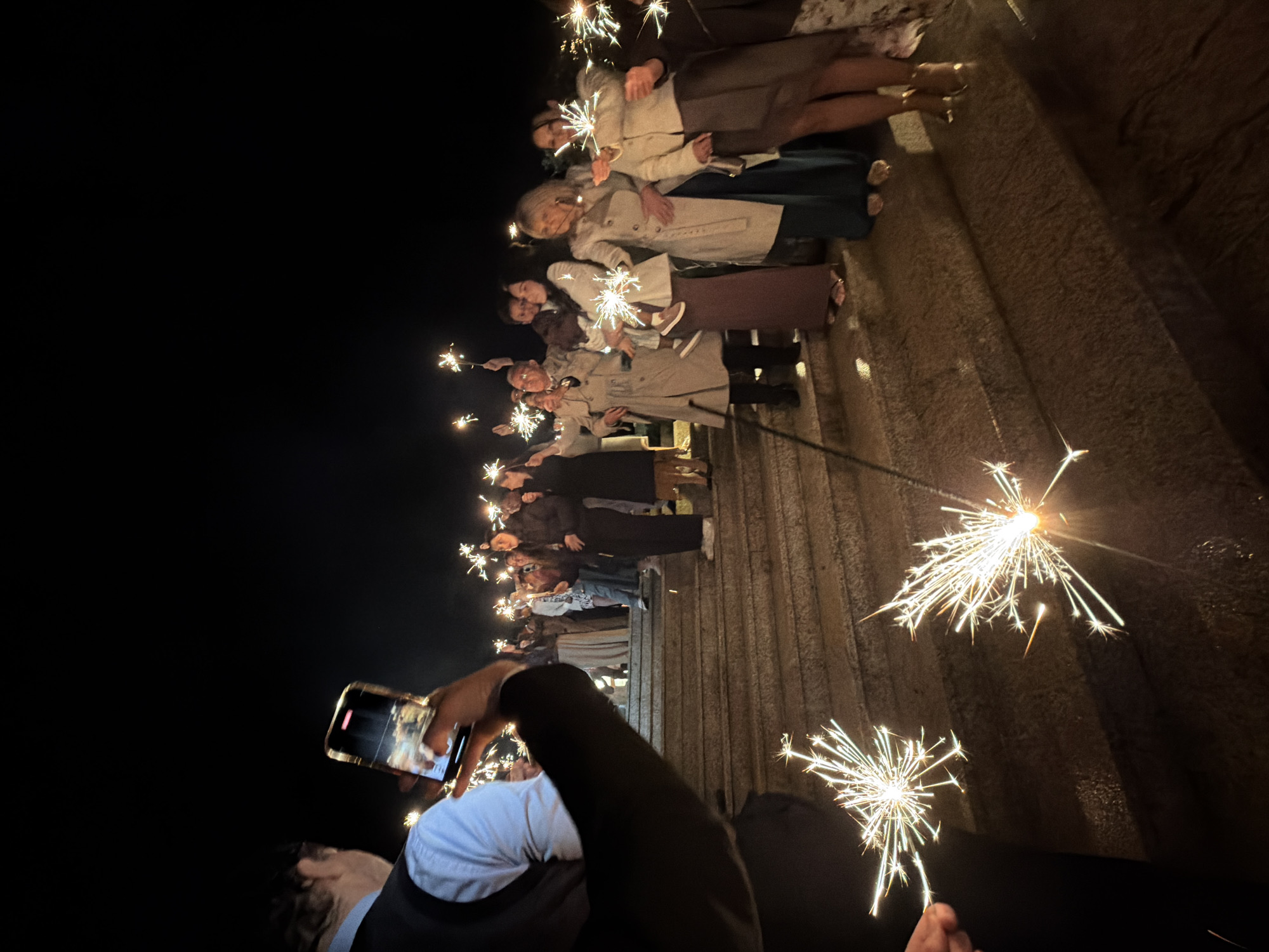 At night in Vila Real, Portugal, a crowd stands in front of an elevated stone structure holding sparklers. A person in the foreground holds up a cell phone to record them. Most of the individuals are wearing coats, and some are holding sparklers. Two large sparklers are in the foreground.