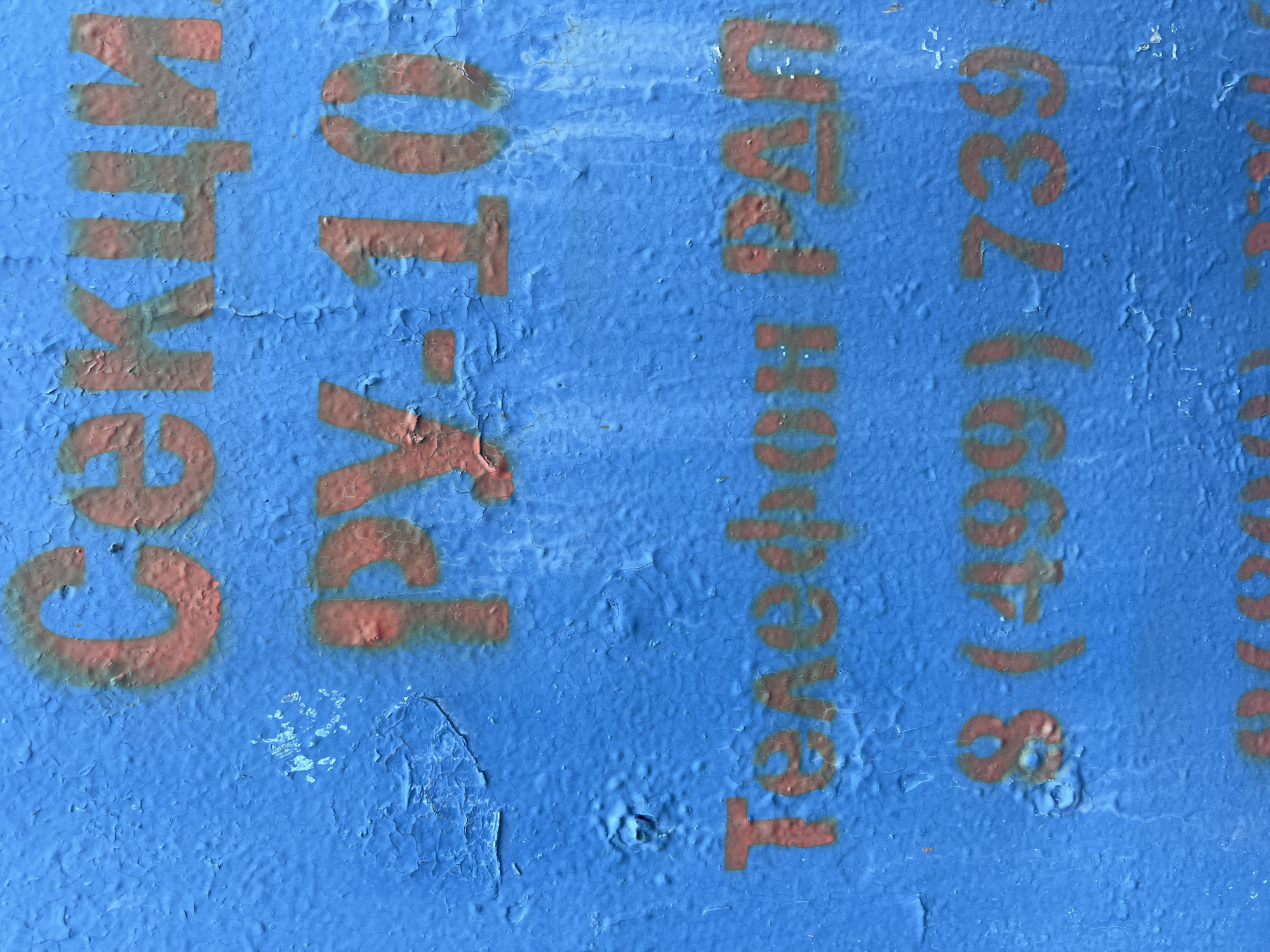 The image shows a blue, weathered wall with stenciled red lettering. The text is in Russian and appears to be related to a section or department. The top line reads "СЕКЦИ" which is likely an abbreviation for "секция" (section). The second line reads "РУ-10", possibly an identifier for the section. The third line says "Телефон РА" which means "Telephone RA". The fourth line is a phone number starting with "8 (499) 739". This phone number format suggests a Moscow area code (499) and likely belongs to a business or organization in Moscow. The blue paint on the wall is cracked and peeling, indicating age and exposure to the elements. There are no people or actions depicted in the image. The scene is a close-up of the text on the wall, with no broader environmental context visible.