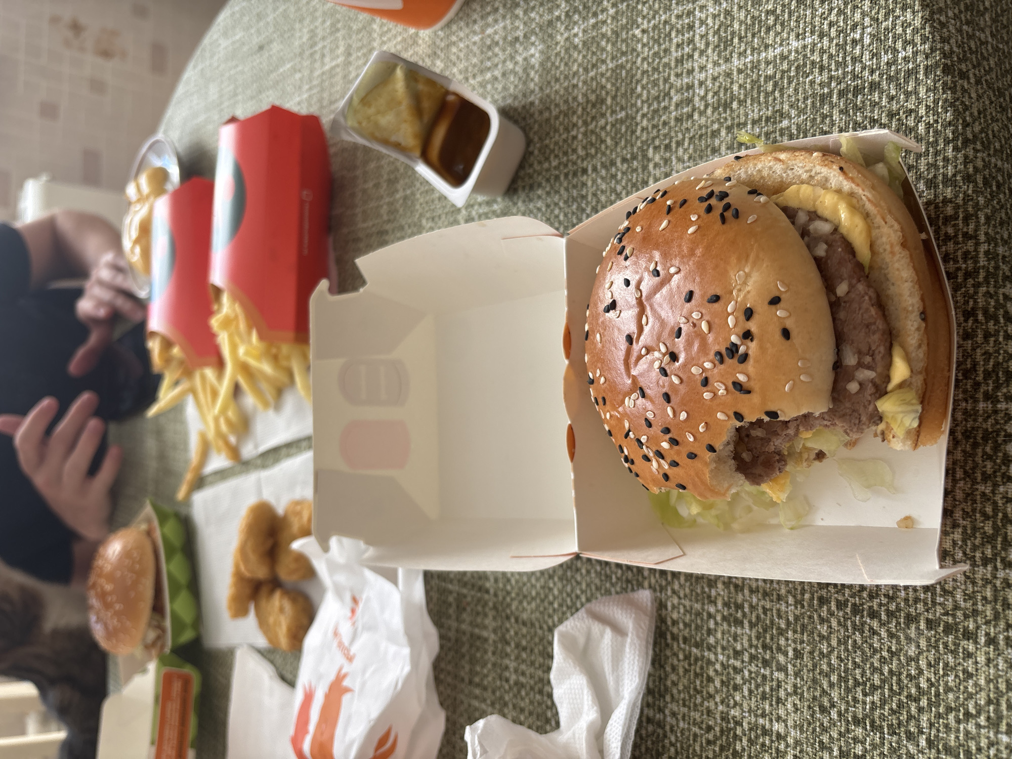 The image shows a close-up view of a partially eaten hamburger and french fries laid out on a table with a green textured tablecloth. The hamburger is in a white cardboard box and has sesame seeds on the bun. There are also chicken nuggets and dipping sauce visible. In the background, two people are partially visible, with one person's hands resting on the table. The setting appears to be an indoor dining area. There are no identifiable landmarks or features to indicate the specific location in the city of Sinyavino, Russia.