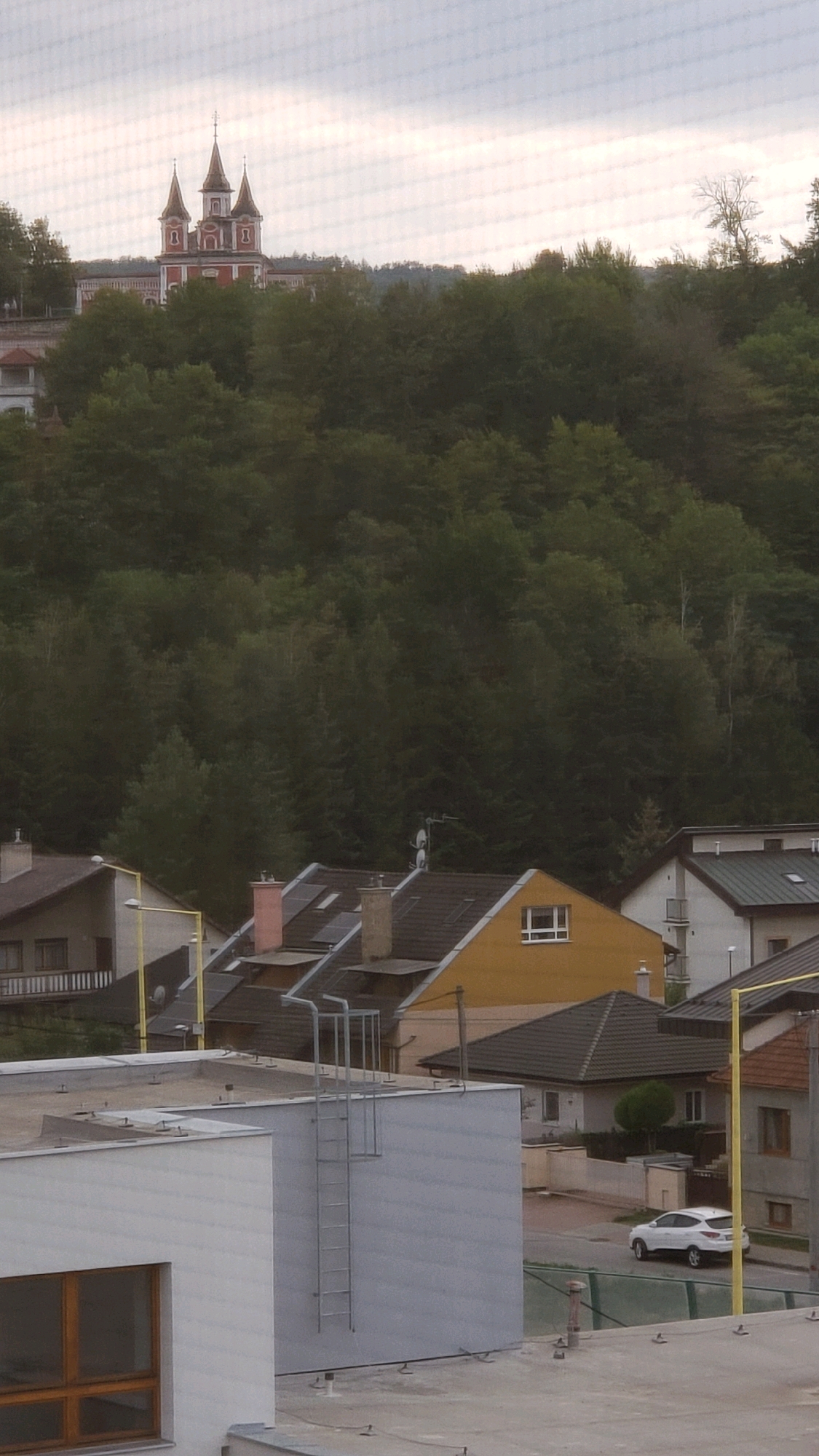 The image shows a residential area in Prešov, Slovakia, on an overcast day. In the background, on a hill covered with trees, a building with multiple red-domed towers and white accents is visible. Below the hill, several houses with sloped roofs and chimneys are clustered together. One house has a bright yellow facade and a white-framed window. In the foreground, a modern building with a flat, light-colored roof and a large window with dark wooden framing is partially visible. A metal ladder extends vertically up the side of this building. A white car is parked on a street to the right of the houses. The overall impression is of a tranquil, suburban neighborhood.