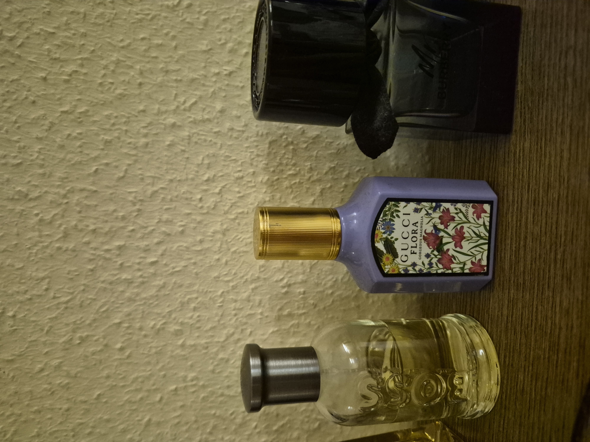 The image shows three perfume bottles on a wooden shelf against a textured, light-colored wall. From left to right, the first bottle is clear glass with a metallic gray cap and contains a light yellow liquid. The second bottle is lavender-colored with a gold cap and a label featuring floral illustrations and the text "GUCCI FLORA GORGEOUS MAGNOLIA". The third bottle is dark glass with a black cap and the visible text "BURBERRY". The lighting suggests indoor conditions, likely in a domestic setting.