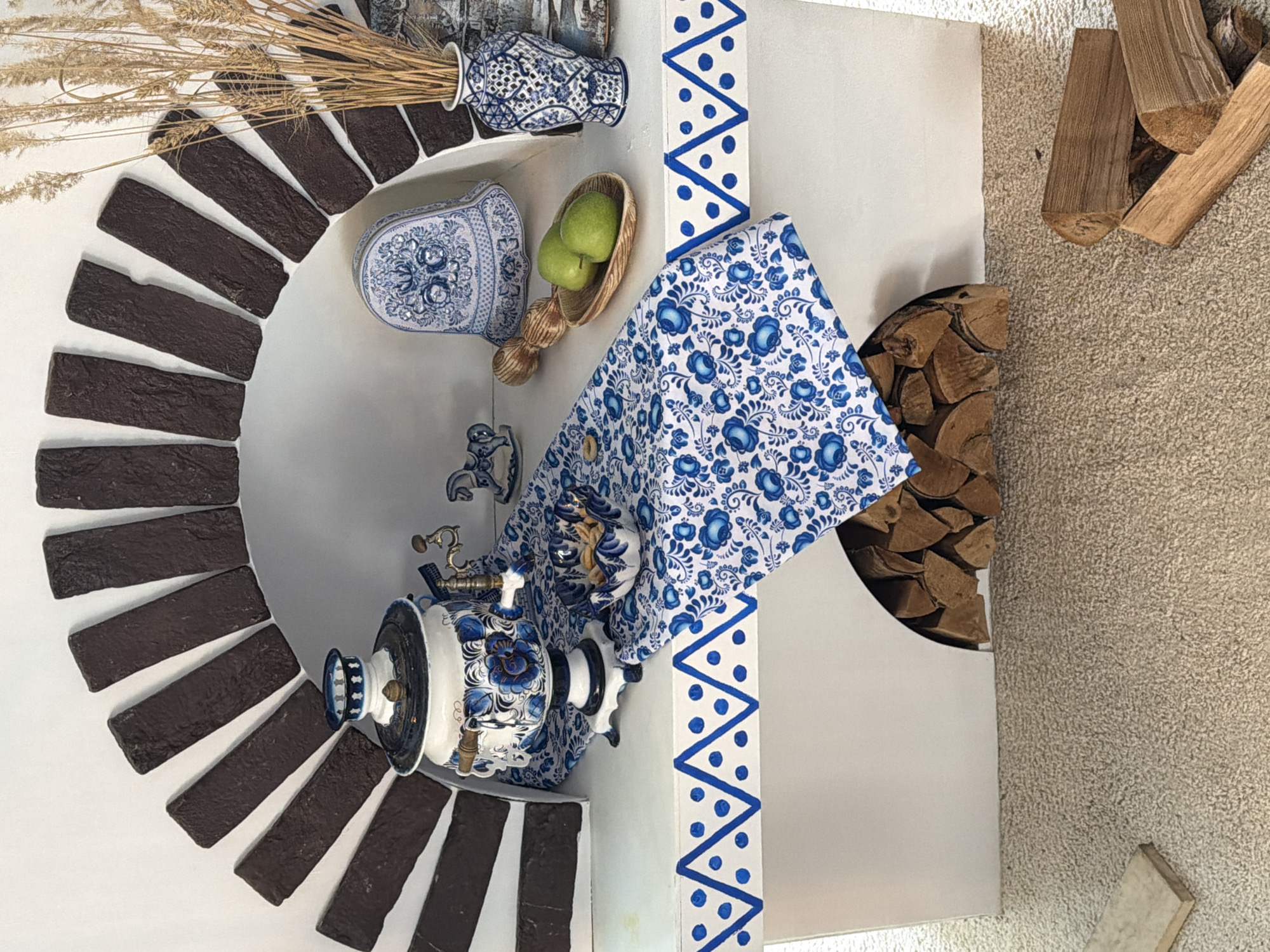The scene is set indoors, within the city of Izhevsk, Russia, displaying a white decorative structure resembling a stylized fireplace unit. The upper section features an arched recess lined with dark brown rectangular bricks. On the flat white surface below this arch, various objects are arranged. From left, a white samovar decorated with blue floral (Gzhel-style) patterns rests on a matching blue and white patterned fabric runner. This runner drapes over the structure's front edge, which is adorned with a continuous blue zigzag and dot motif. To the right of the samovar, a small, similarly patterned bowl holds light-colored nuts. Further right are a small ceramic rocking horse with blue details, a white bell-shaped decorative item with blue patterns, and a wooden bowl containing two green apples. A blue and white patterned vase filled with dried wheat stalks stands on the far right, in front of a partially visible background image. Below the decorated edge, a lower arched recess holds neatly stacked firewood. The floor is covered with light beige carpet, upon which several additional pieces of cut firewood are scattered in the foreground. No individuals are present in the image.