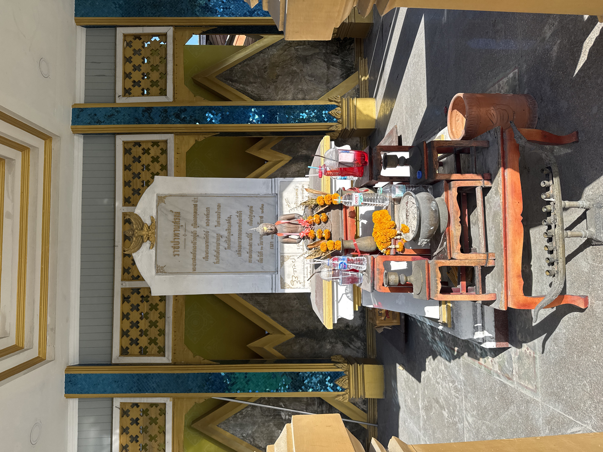 The image depicts an outdoor shrine likely located in Patong, Thailand, possibly near a temple or a place of cultural significance. In the center of the frame, a stone stele with Thai inscriptions stands prominently, topped with a Garuda emblem. In front of the stele, a small, weathered statue of a seated Buddha is adorned with orange marigold garlands.
Arranged before the Buddha statue is an altar set on tiered wooden stands painted red and orange. On the altar, various offerings are present, including small bottles of water, plastic cups with red liquid, and a censer containing ashes. Below the altar, on the ground, are additional offerings and decorative items, including a brass candelabra with candle remnants and a terracotta pot.
The shrine is situated in an open paved area, with shadows cast by overhead structures indicating a sunny day. The background features architectural elements with gold and teal decorative panels, suggestive of traditional Thai design. The overall scene conveys a sense of religious devotion and cultural tradition.