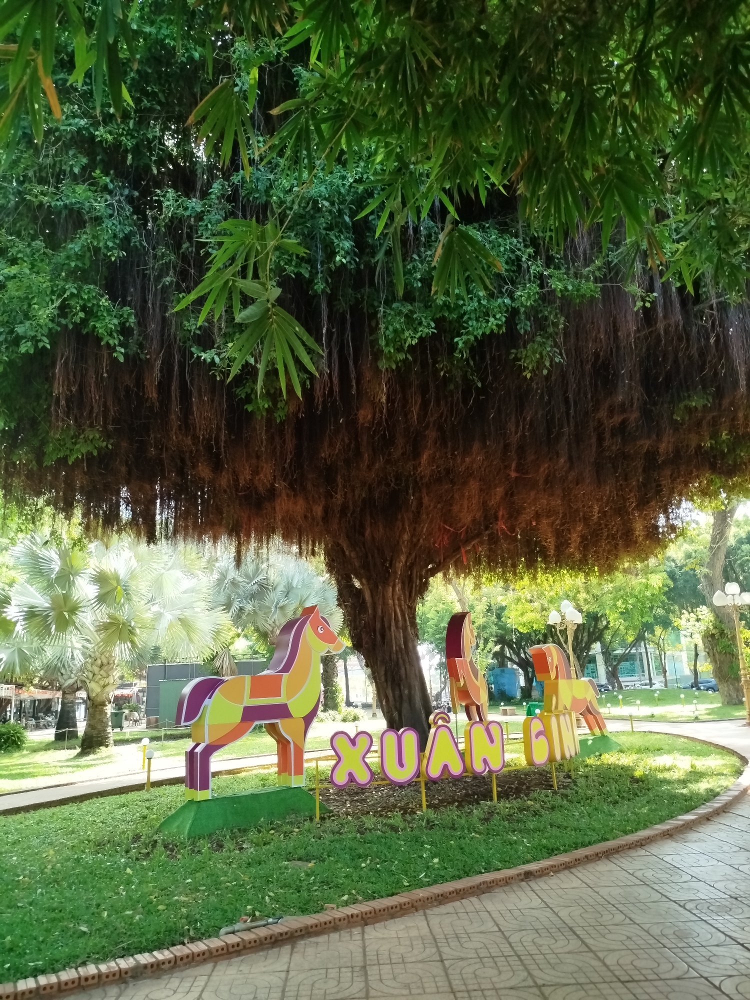 The image shows a park scene in Ho Chi Minh City, Vietnam, featuring a large banyan tree with numerous aerial roots hanging down. Beneath the tree are colorful cutout horse figures and the word "XUAN," which translates to "Spring" in Vietnamese, in pink and yellow letters. The area around the figures is a small patch of green grass. The park has other trees and greenery in the background. The foreground includes a paved walkway made of gray and beige tiles.