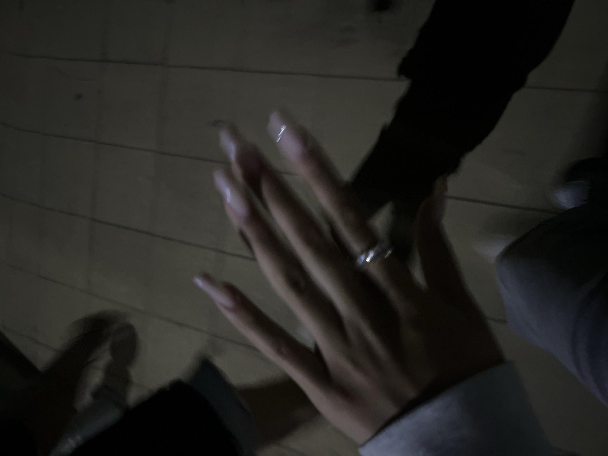 The image depicts a low-light, blurry scene in an unknown location within the city of Unknown, Unknown. The primary subject is a human left hand, positioned centrally and slightly to the left, extended forward with fingers splayed. The hand has long, light-colored fingernails, possibly coffin or almond shaped, with a reflective quality on the tips. A silver-toned ring, appearing to have small stones, is worn on the ring finger. The wrist is partially covered by a light grey or blue-grey long sleeve.
The background consists of a tiled or panelled floor with dark grout lines, rendered in muted grey and brown tones due to the dim lighting. In the lower left, dark fabric, consistent with trousers or jeans, is visible on what appears to be the lap area of the person whose hand is shown, with a faint light reflection on its surface. Additional blurry, dark shapes, possibly an arm or clothing of another individual, are present in the upper left background. A prominent, dark shadow is cast to the right of the main hand. The overall image quality exhibits significant motion blur, consistent with a moving subject or low-light conditions and a slower shutter speed.