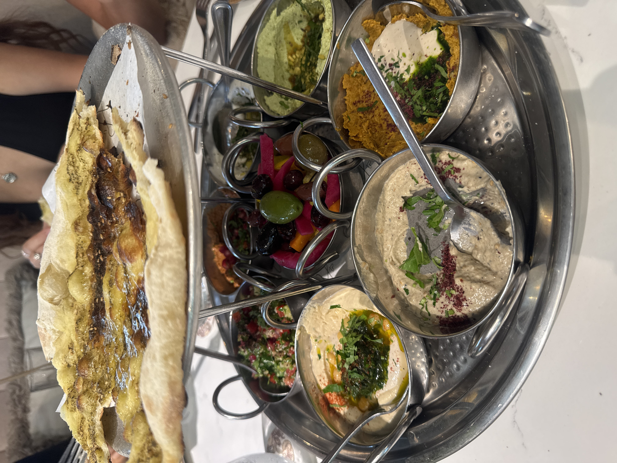 A tiered metal platter holding various dips and bread is displayed on a white marble surface in a restaurant in Aventura, Florida. The platter's top tier contains a large piece of flatbread topped with what appears to be za'atar and olive oil. The middle tier holds several small metal bowls filled with assorted dips, including a vibrant green dip, a reddish-orange dip with pomegranate seeds, a mixture of olives and pickled vegetables, and a colorful chopped salad. The bottom tier of the platter features additional dips, one with a creamy texture and green garnish, and another a lighter colored dip garnished with sumac and parsley. Spoons are present in several of the bowls. In the background, a person wearing a black top is partially visible. The lighting is bright, suggesting an indoor setting.