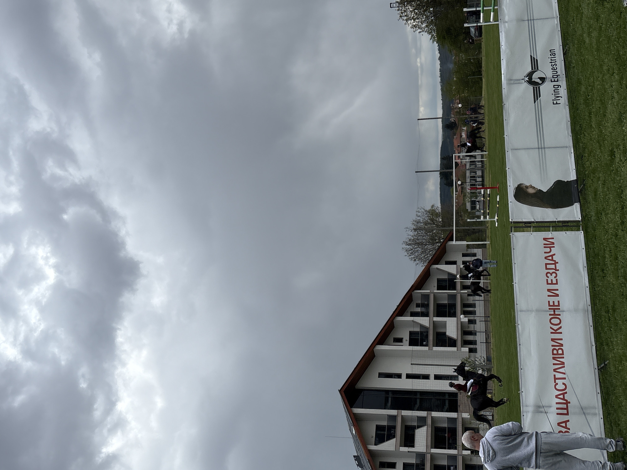 The image depicts an equestrian event in Panagyurishte, Bulgaria, under a cloudy sky. In the foreground, banners display the logo "Flying Equestrian" and Cyrillic text. A man in a grey sweatsuit is visible at the bottom. Horses and riders are seen jumping over obstacles in the background. A modern building with a distinct roofline is also present. A net surrounds the field with spectators watching.