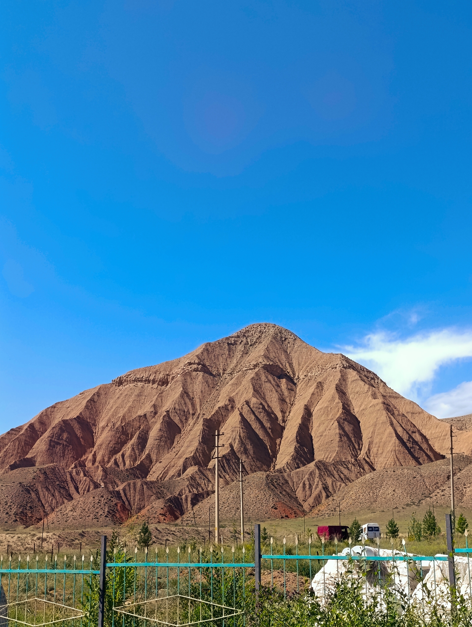 The image depicts a large, eroded mountain range with sharp, jagged peaks, likely composed of sedimentary rock, under a clear blue sky. The mountains display a reddish-brown hue with visible striations and gullies, suggesting significant weathering and erosion. In the foreground, a light green metal fence with pointed finials runs horizontally across the frame. Behind the fence, there is dry, sparse vegetation, with some scattered green bushes and a few small trees. Two utility poles are visible in the middle ground, one closer to the right side of the frame, and another further back towards the center. In the distance, behind the fence and some trees, two vehicles are present: a red van and a white van or bus. A few white objects, possibly bags or tarps, are also visible in the lower right portion of the image. The overall scene suggests a semi-arid or mountainous region. The location is identified as the city of Naryn, Kyrgyzstan.