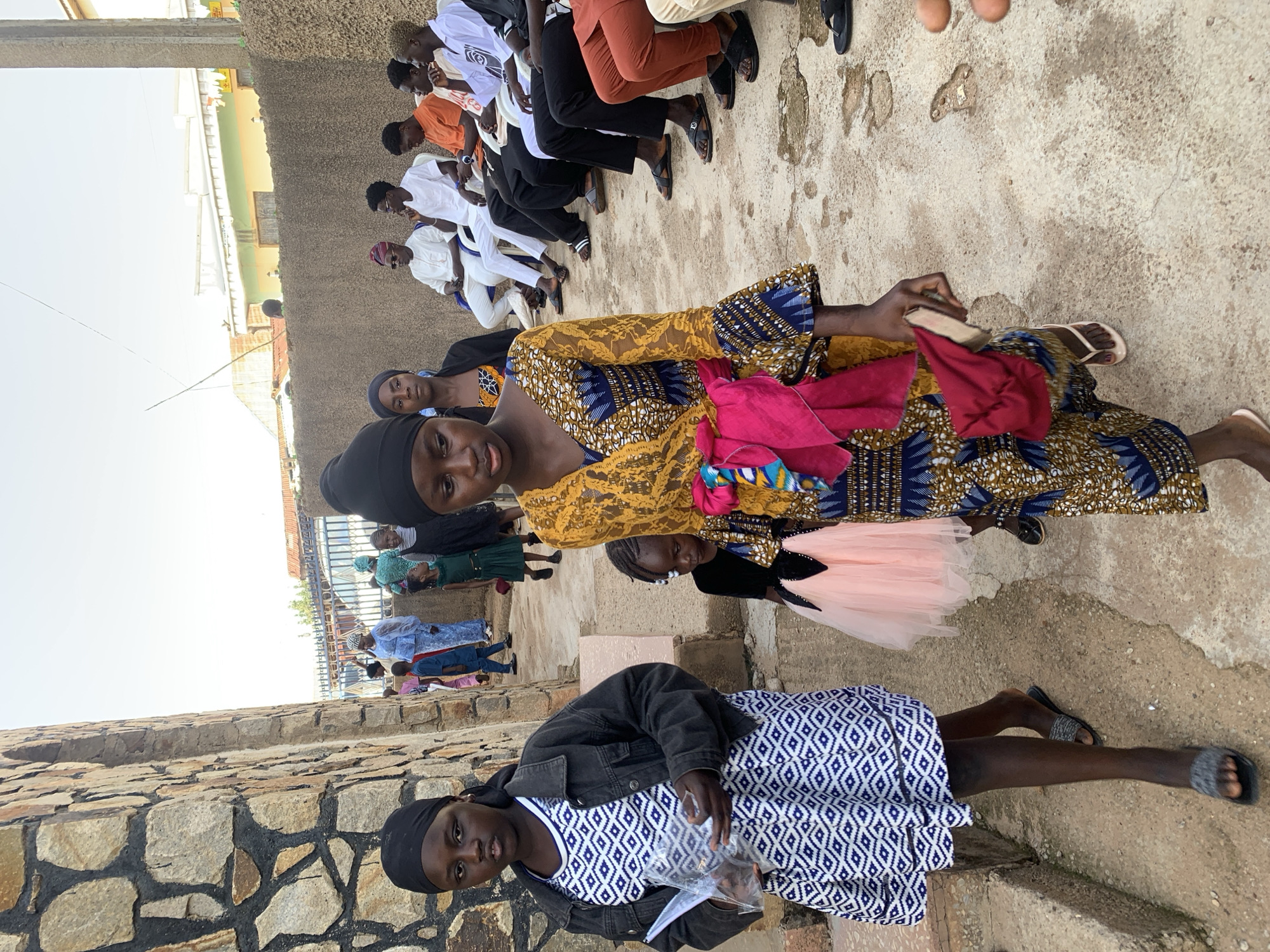 A group of people is outdoors in what appears to be a gathering or event. In the foreground, two young women are prominent, both wearing head coverings. One is dressed in a blue and white patterned dress with a denim jacket, holding a clear plastic bag. The other wears a gold and blue patterned dress and carries colorful cloth items. Several other people, including children in dresses, are seated or standing in the background, along a concrete or stone wall. Further in the background, men in various attire are also seated. The setting appears to be urban, with buildings visible in the distance. The location is given as Jos, Nigeria, a city on the Jos Plateau in the Middle Belt of Nigeria.