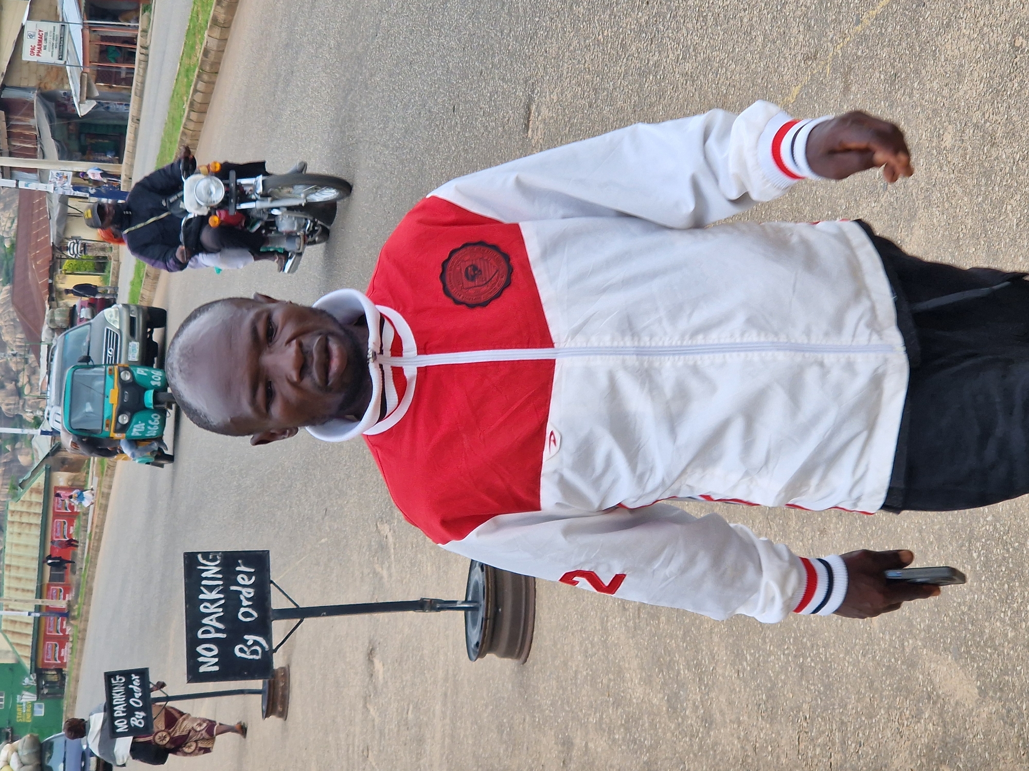 In Kabong, Nigeria, an outdoor scene unfolds on a paved road. A man, viewed from the chest up, walks towards the camera, holding a smartphone in his right hand. He is wearing a white and red jacket with a circular emblem on the left chest and red trim on the cuffs. His left sleeve features a red number "2". In the background, a woman walks away from the camera, carrying a bag over her left shoulder and wearing a patterned skirt and a grey shawl. Two "NO PARKING By Order" signs are visible on the sidewalk. A teal and yellow auto-rickshaw, labeled "PTA 3660", and a silver SUV are on the road. A man on a red motorcycle, wearing a black jacket and cap, is also on the road, with a passenger behind him. Buildings with red roofs and green trim line the right side of the street. The overall environment appears to be a moderately busy street with pedestrian and vehicular traffic.