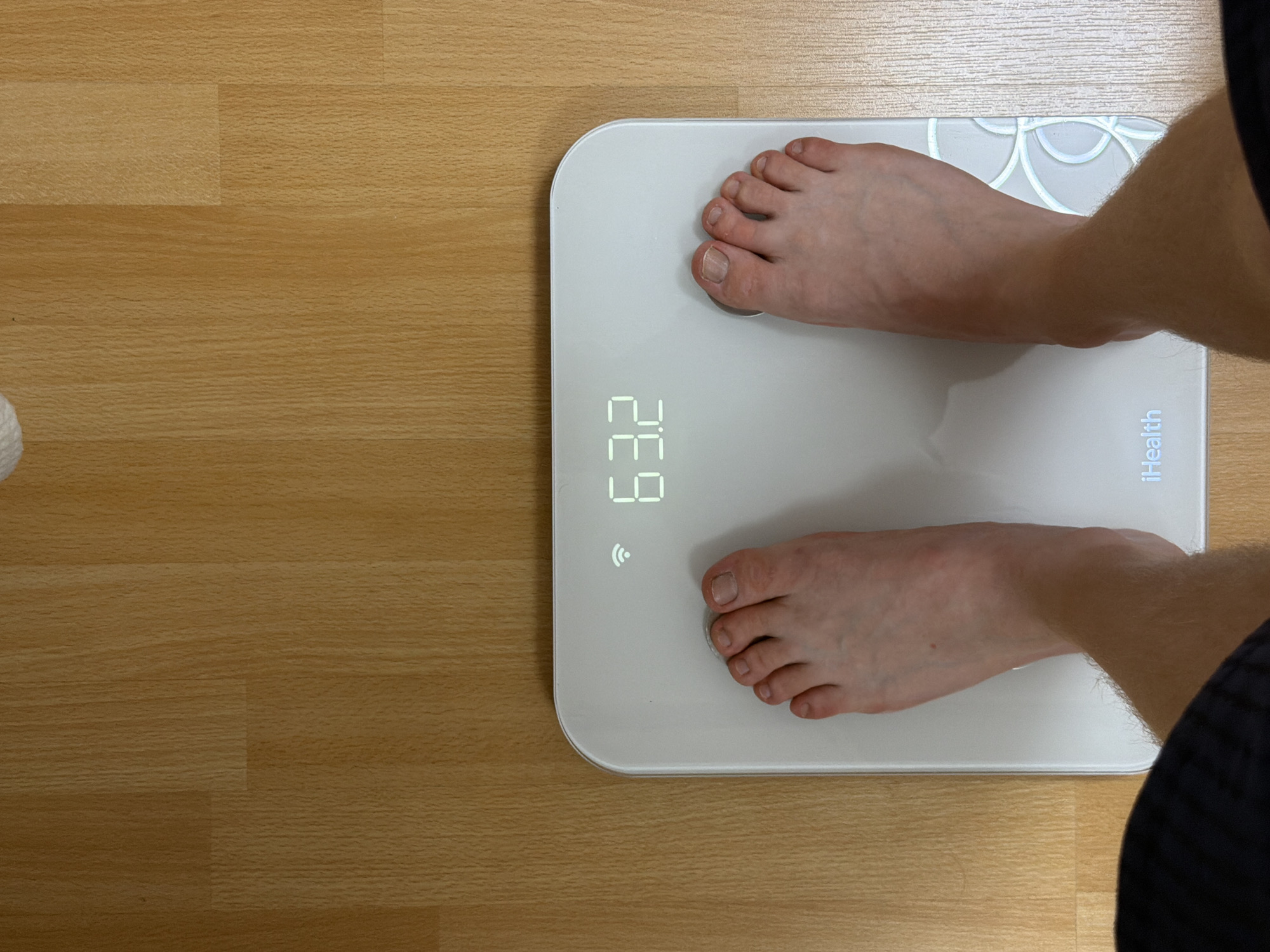 A person is standing on a white iHealth smart scale on a light wood floor. The scale displays the weight "63.2" in glowing digits. The person's bare feet are positioned on the scale, with their toes visible. The background is a wooden floor with visible grain patterns. The image is taken from a top-down perspective.