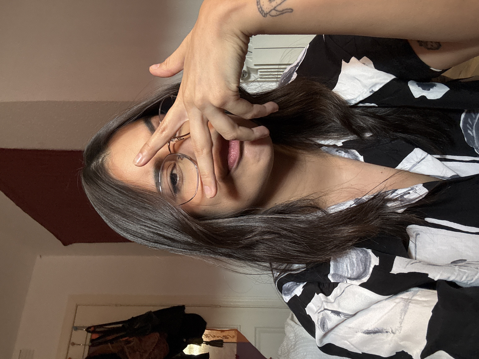 A young woman with long, dark hair and glasses is posing for a selfie. She is wearing a black and white floral print shirt. Her right hand is covering part of her face, with her fingers splayed. There is a tattoo on her left forearm. The background appears to be a bedroom, with a closet and a bed visible. The lighting is dim. There is no information available to confirm or deny the location in Las Rozas de Madrid, Spain.