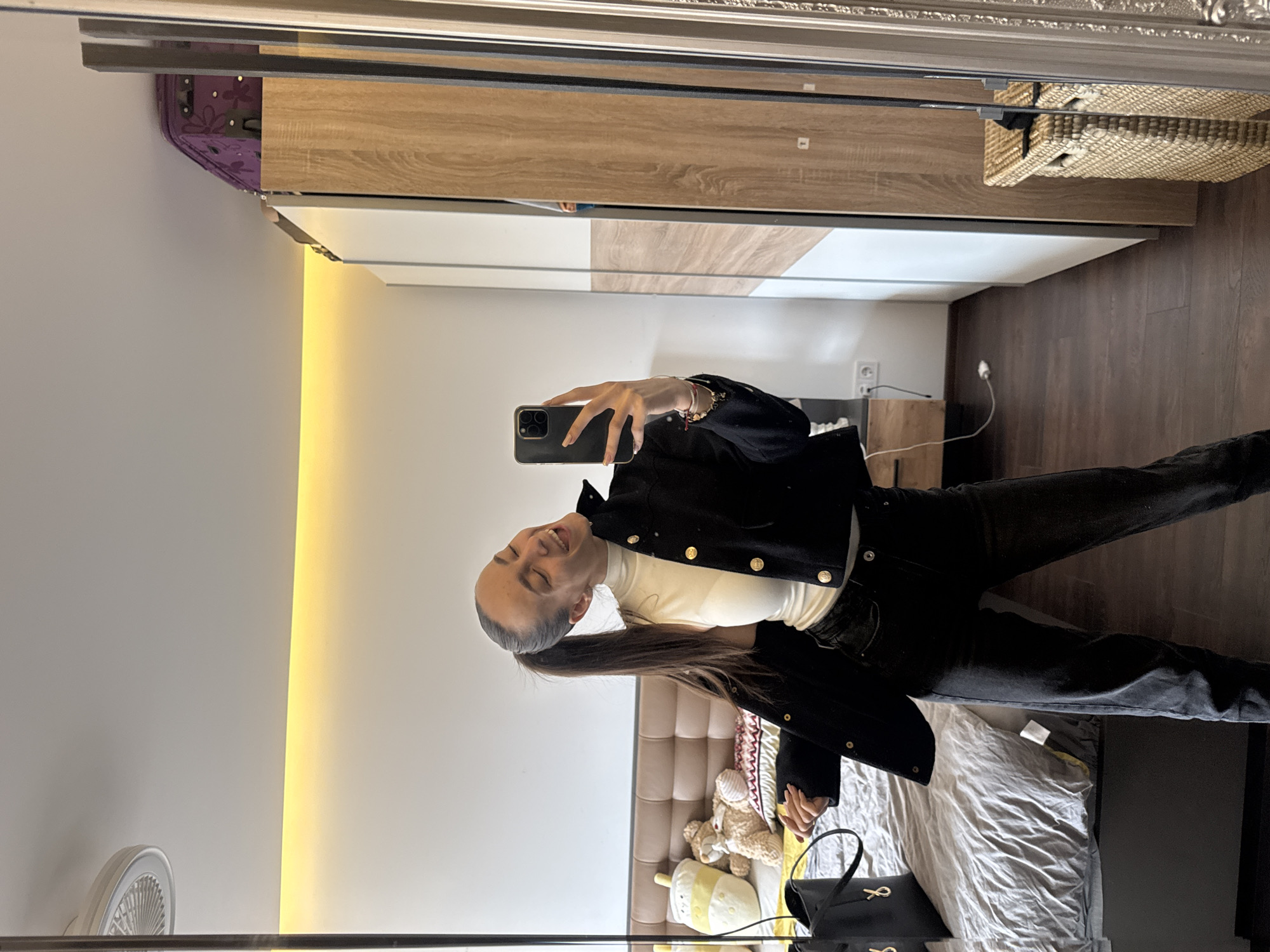 A person is taking a selfie in a mirror. The person is wearing a white top, black jacket and black pants. The background appears to be a bedroom with a bed, wardrobe, and bedside table. The city and country are irrelevant to the image's content.