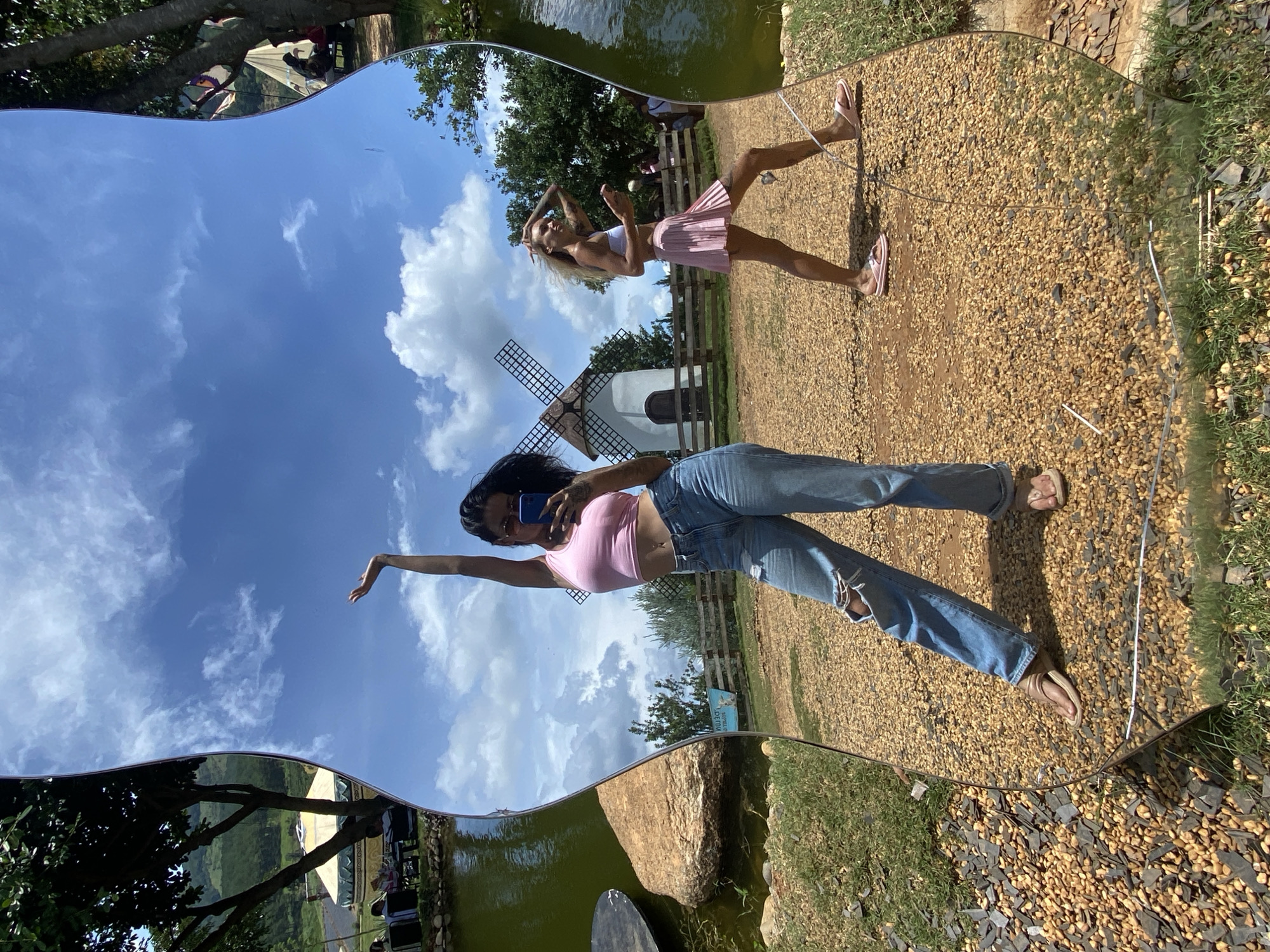 A full-body mirror, shaped like a guitar, reflects two young women posing in an outdoor setting with a windmill in the background. The woman on the left, wearing a pink crop top and ripped denim jeans, stands facing the mirror, holding a blue smartphone and raising her left arm. Her right arm is bent at the elbow with her hand near her hip. The woman on the right, dressed in a white crop top and a pink pleated skirt, is captured in mid-stride, looking to her right and touching her hair. She wears pink flip-flops. The mirrored surface reflects a clear blue sky with fluffy white clouds, a green body of water on the left, trees, and a white windmill with a brown roof and black blades situated behind a wooden fence. The ground is covered in small pebbles and scattered green grass. In the distance, obscured by the mirror's reflection, there are tents and people seated at tables. The overall scene depicts a sunny day at what appears to be a recreational area or park. The location is Thôn 5, Vietnam.