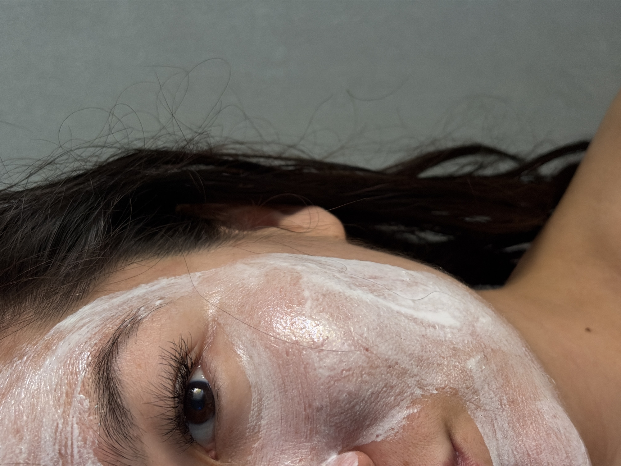 This is a close-up, right-side profile shot of a person's face, partially covered in a white cream or mask. The subject has dark hair, and their right eye is visible. The cream is applied unevenly across the cheek and forehead. The background is a plain, light gray wall. The image does not contain any elements that can be used to determine its location in Chaikovsky, Russia.