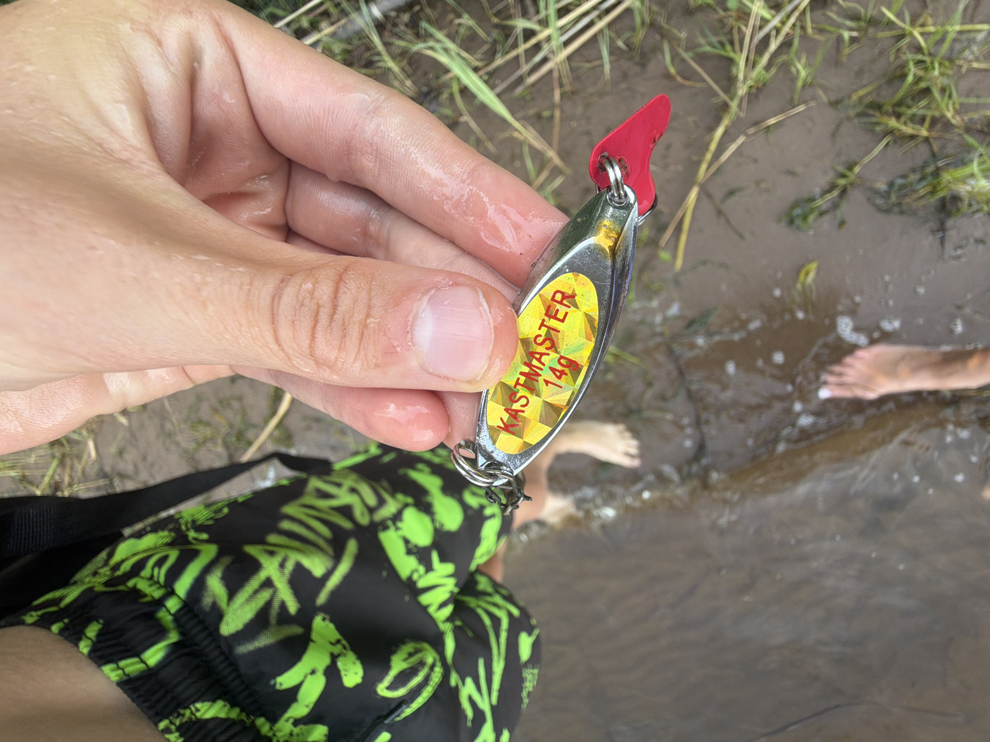 The image shows a close-up of a hand holding a fishing lure. The lure is silver with a yellow and red checkered pattern and the words "KASTMASTER 14G" printed on it in red letters. The hand is wet, and there are water droplets on the skin. In the background, a person's legs are visible, wearing black shorts with neon green patterns. The person is standing in shallow, murky water with some green grass growing along the bank. The overall scene suggests an outdoor setting, likely near a body of water, possibly a river or lake. The specific location in Chaykovsky, Russia cannot be determined from the image alone.