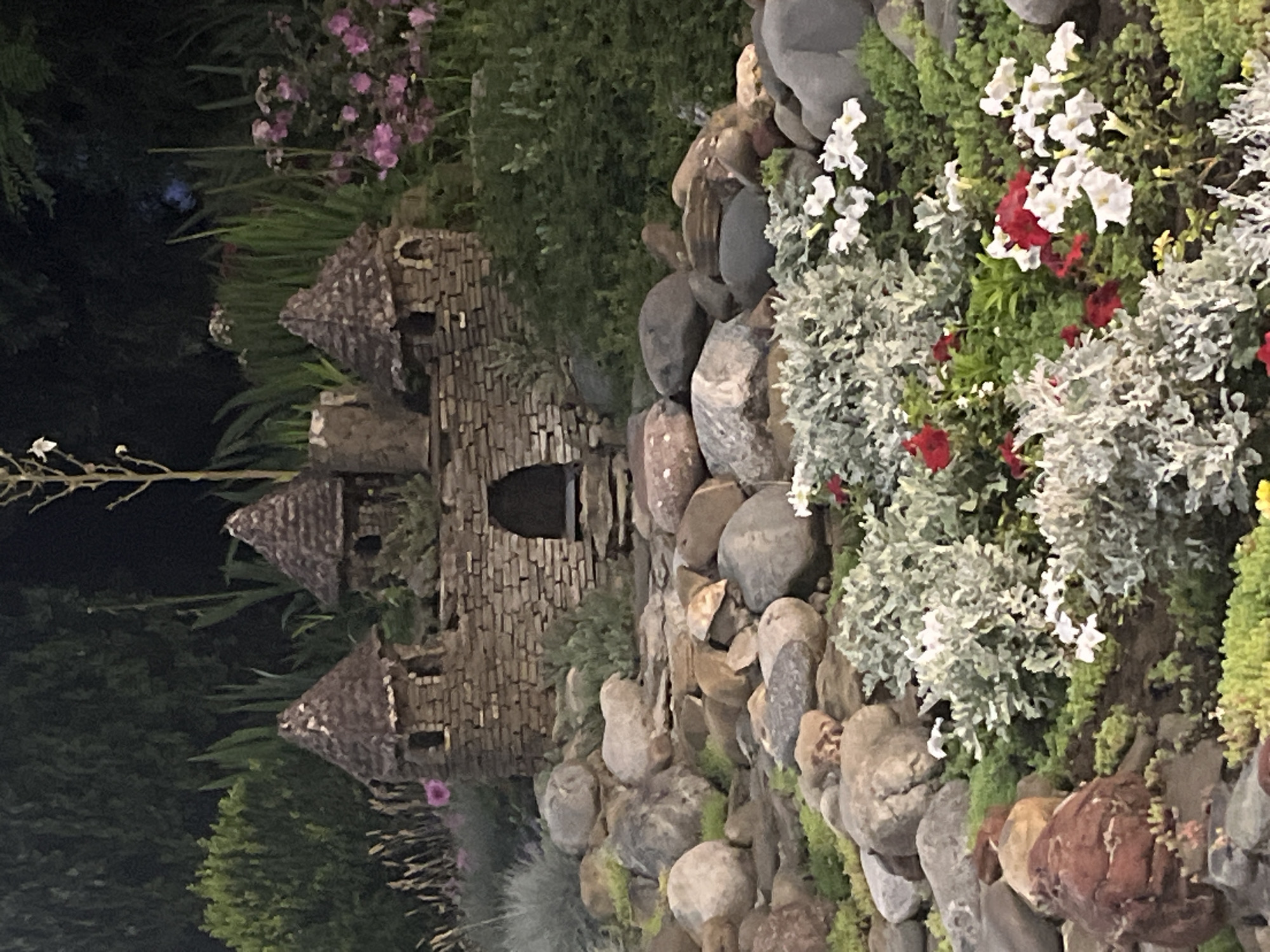 The image depicts a miniature stone castle nestled in a garden landscape. The castle is constructed from rough-hewn stones, featuring three conical turrets and a central arched entryway with visible steps. It is surrounded by a rock garden with various sizes of stones, some appearing moss-covered. In the foreground, there are clusters of low-lying plants, some with silvery-green foliage and small white and red flowers. Behind the castle, a dense arrangement of dark green foliage and taller plants creates a backdrop. The lighting suggests it is either dusk or a shaded area, with the dark background hinting at deep shade or possibly the night sky. There are no people visible in the scene. The location is identified as the city of Krasnodar, Russia.
