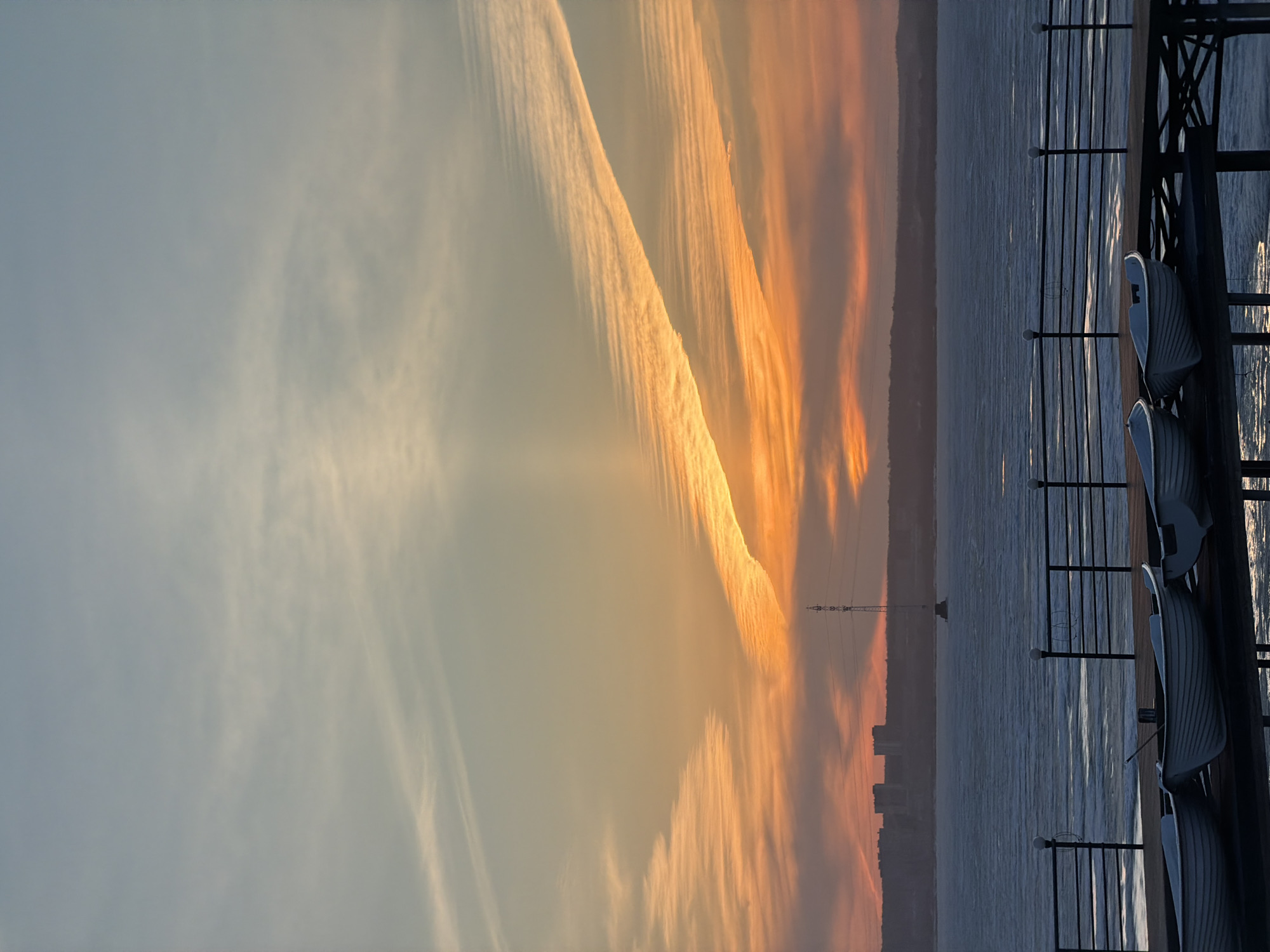 The image shows a sunset view from a pier in Izhevsk, Russia, overlooking the Izhevsk Reservoir. The sky is filled with orange and yellow hues caused by the setting sun and cloud formations. A linear contrail stretches across the sky. A transmission tower and distant buildings are visible on the horizon. The water of the reservoir reflects the colors of the sky. In the foreground, a section of the pier is visible, including a railing and several white chairs.