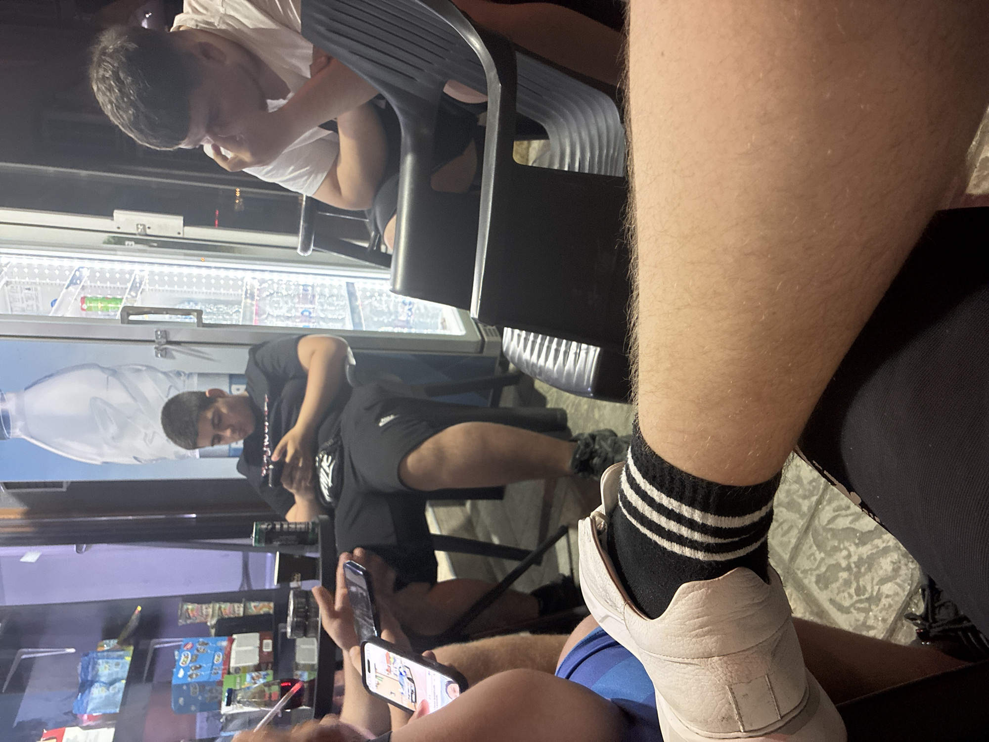 The image depicts a group of people sitting at a table outside what appears to be a vending machine kiosk. One person is looking at their phone. Another person is leaning on their hand. A third person is looking at something off camera. The location appears to be Burgas, Bulgaria, based on the style of vending machine and the streetscape.