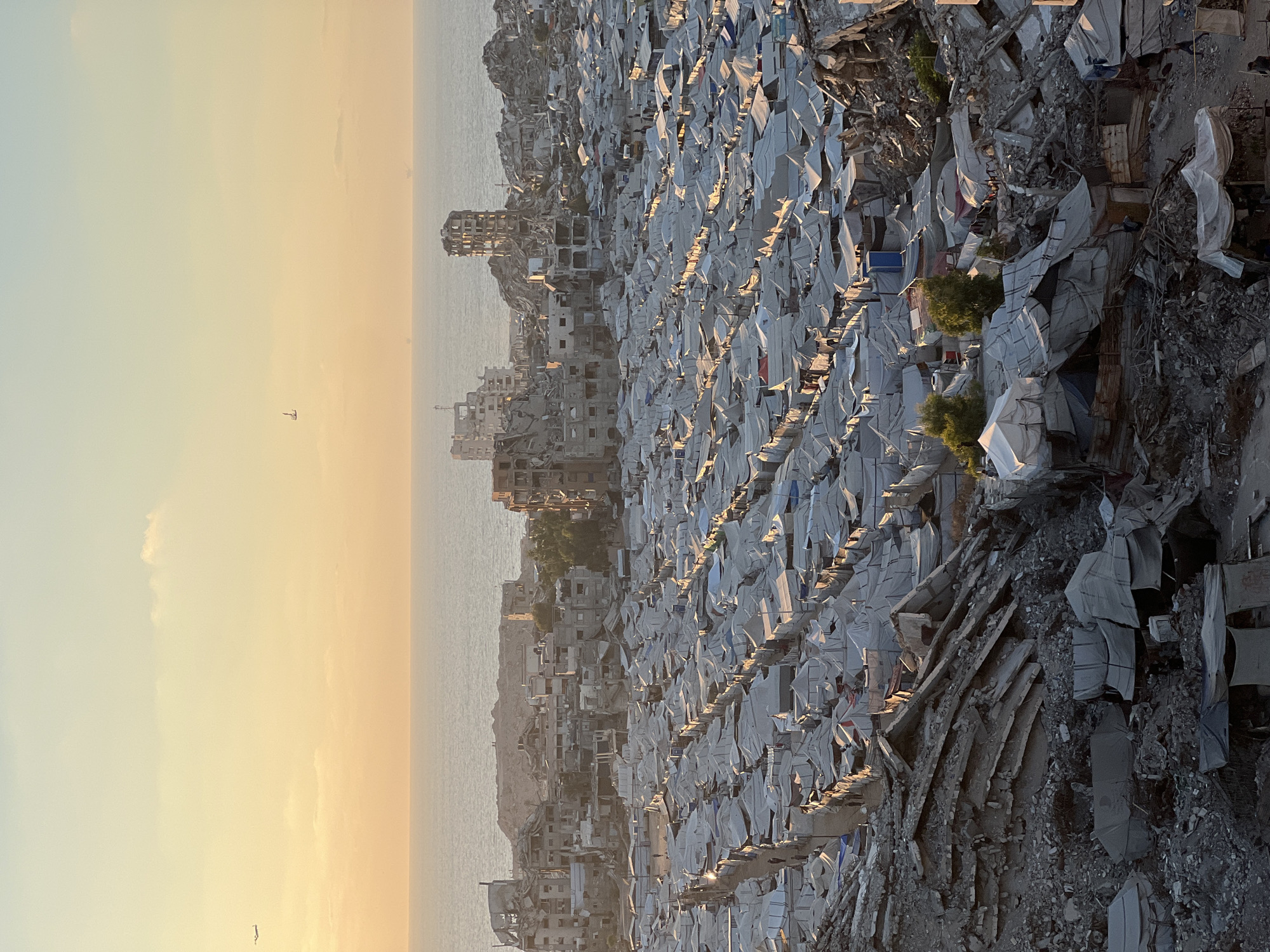 An aerial, high-angle, long shot captures an expansive refugee camp in the city of Gaza, Palestinian Territories. The camp is set against the backdrop of the Mediterranean Sea, with the sun setting and casting a warm, orange glow across the sky and water. In the distance, the remnants of bombed-out buildings stand as stark silhouettes against the horizon, some with their internal structures exposed.
The foreground and middle ground of the image are dominated by a densely packed settlement of white canvas tents, arranged in neat rows and pathways. These tents appear to be temporary shelters, likely erected in the aftermath of destruction. Among the tents, various makeshift structures constructed from salvaged materials are visible, indicating resourcefulness and adaptation in challenging circumstances. Small groups of people can be seen walking between the tents, suggesting the daily life within the camp. The ground is a mix of dirt and rubble, reflecting the damaged urban environment.
Two airplanes are visible in the sky. One is a small plane, further away, while the other is closer and appears larger. The overall impression is one of displacement and resilience, with the vastness of the sea and the setting sun offering a sense of scale and a moment of quiet in the midst of devastation.
