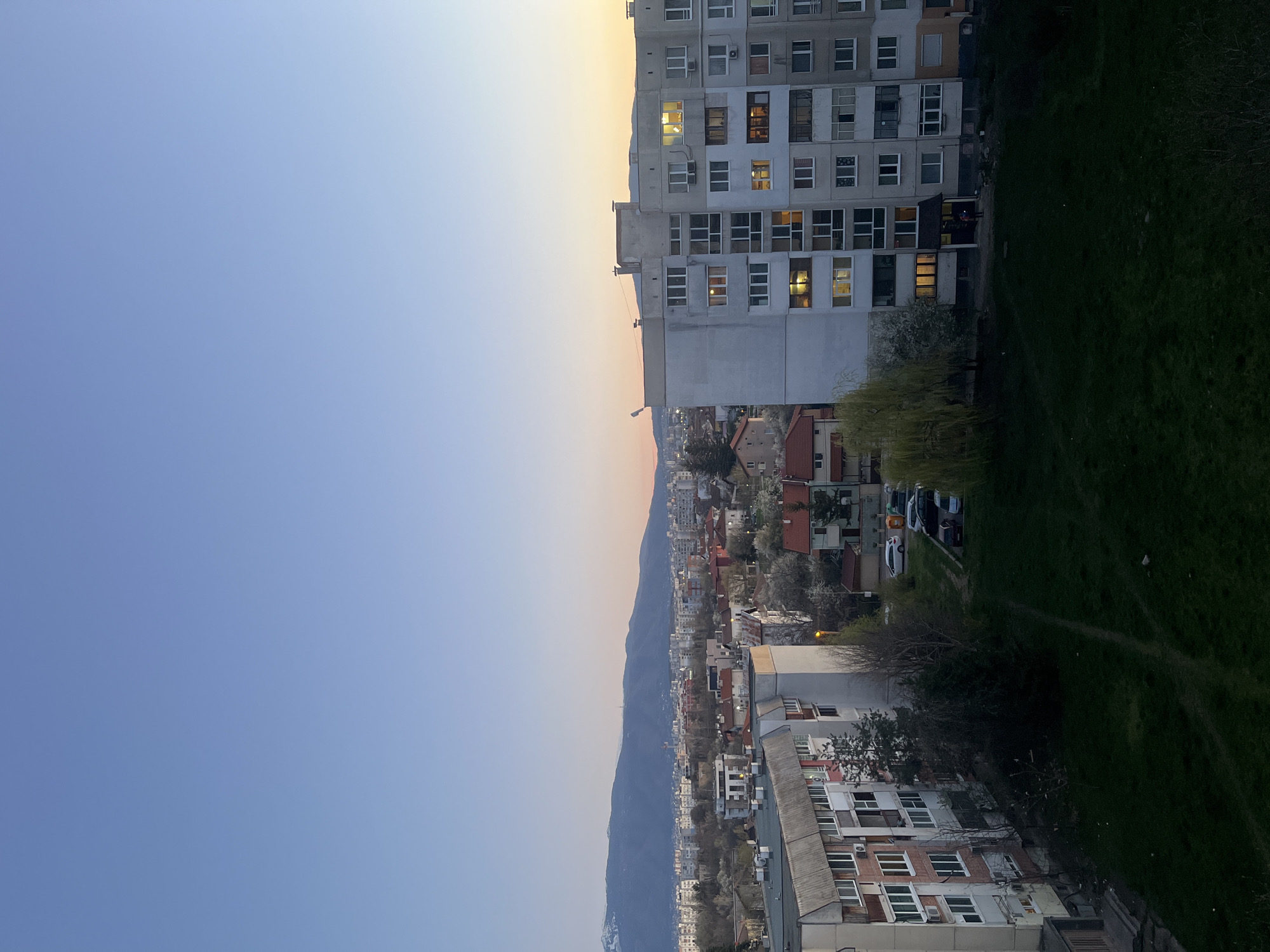 A high-angle, rotated view captures a cityscape, likely in Sofia, Bulgaria, during twilight. A large, grey, Soviet-era apartment building with lit windows is centrally positioned on the right, adjoining a green lawn. The cityscape features various smaller buildings and homes, nestled among trees and vegetation, leading towards a mountain range that bisects the image horizontally. The sky transitions from blue to a light orange/yellow near the horizon. No people are clearly visible, but the lit windows suggest occupancy. Several cars are parked along a road beside the apartment building.